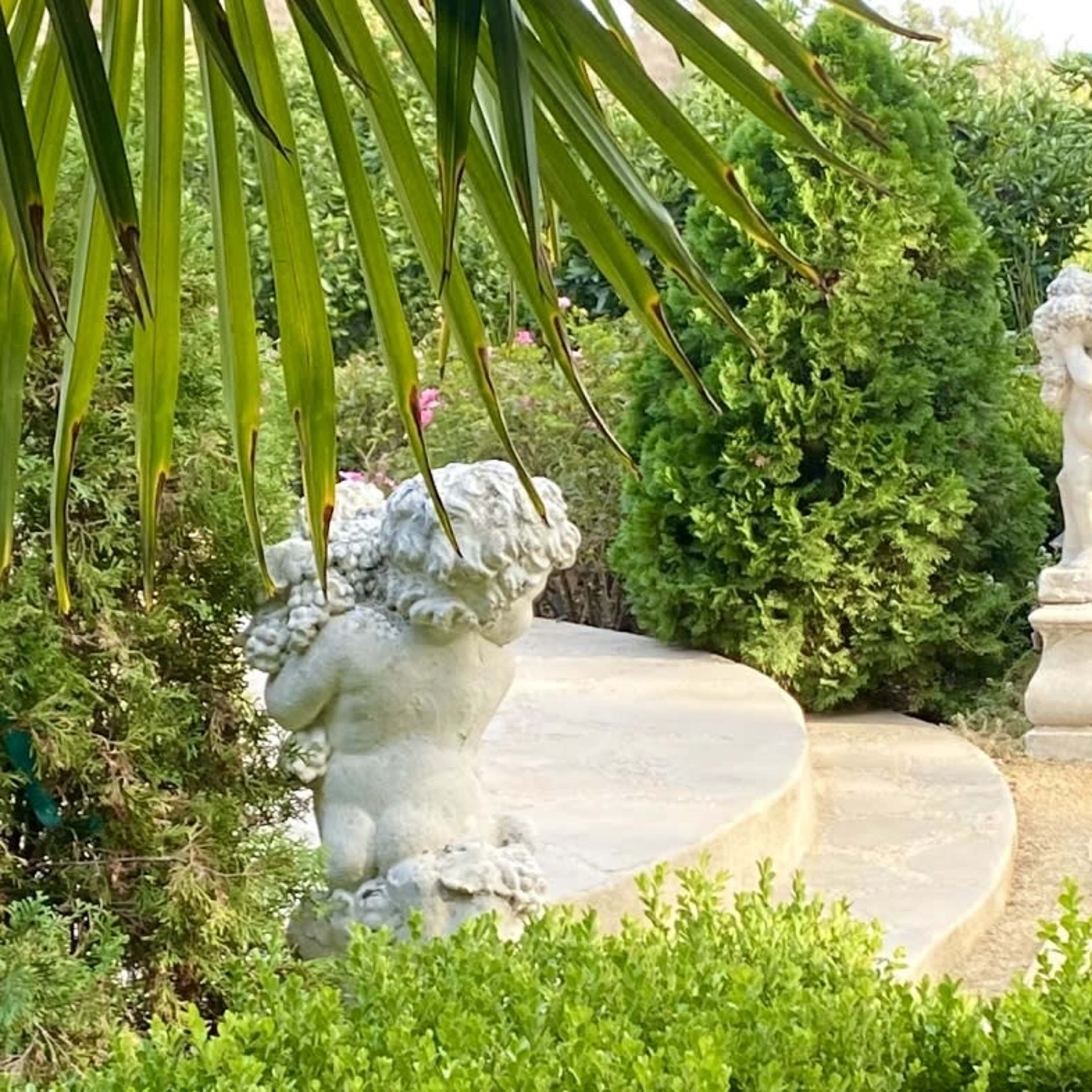 Two stone statues of cherubs positioned in a lush garden, with greenery surrounding a curved pathway.
