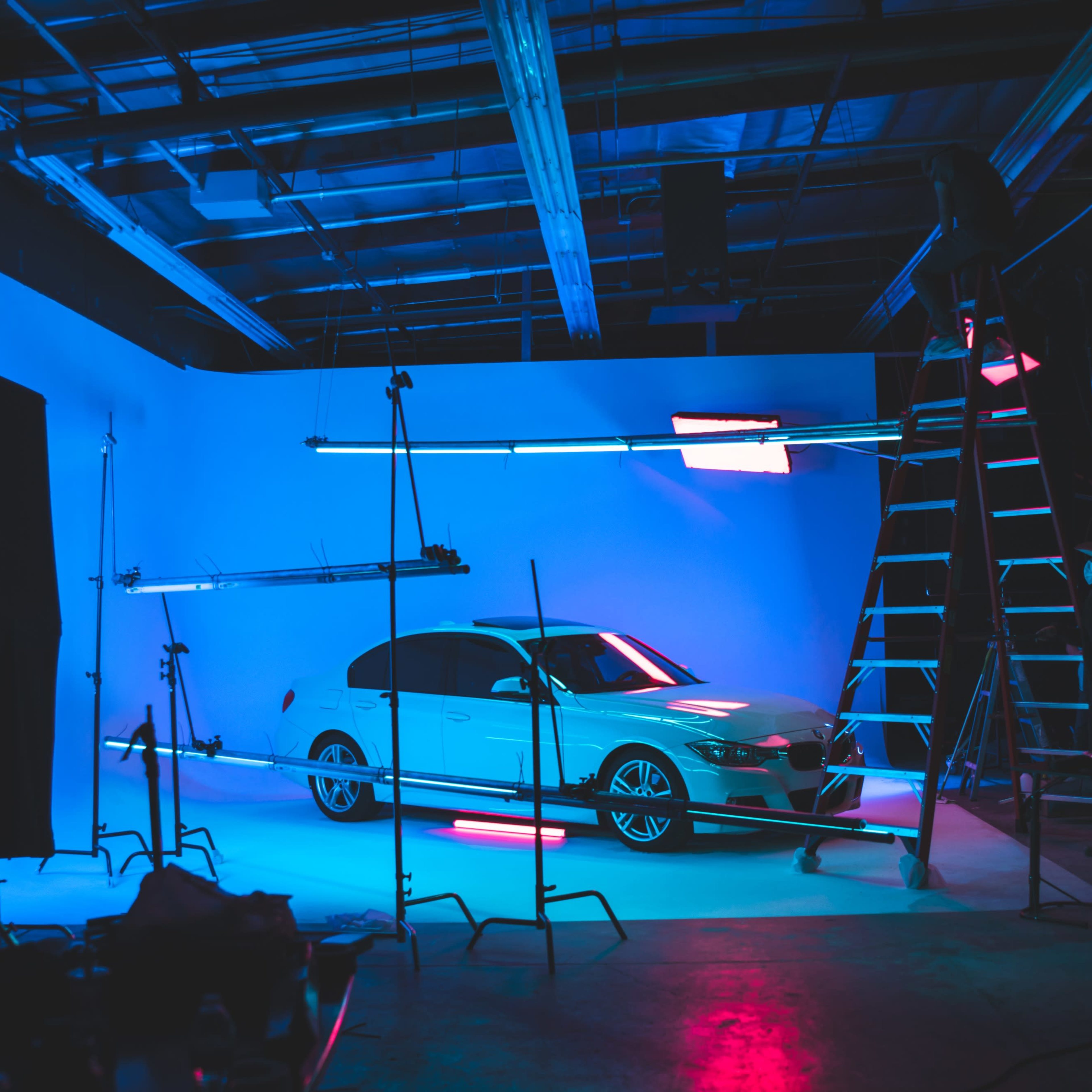 A white car is positioned in a photography studio with blue and pink lighting, surrounded by equipment and a ladder.
