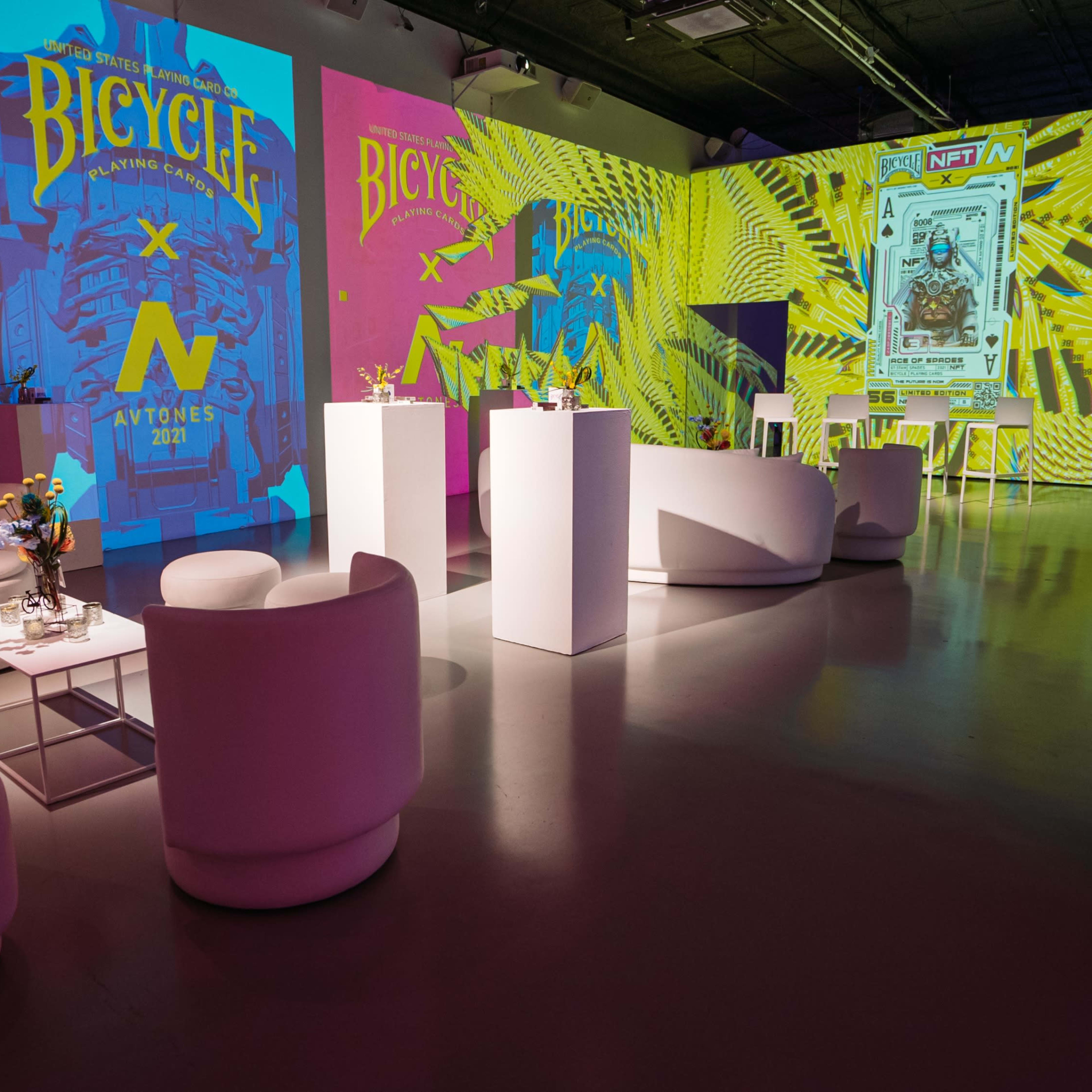 A modern event space with white furniture and vibrant, colorful projections on the walls featuring designs related to bicycles and NFTs.