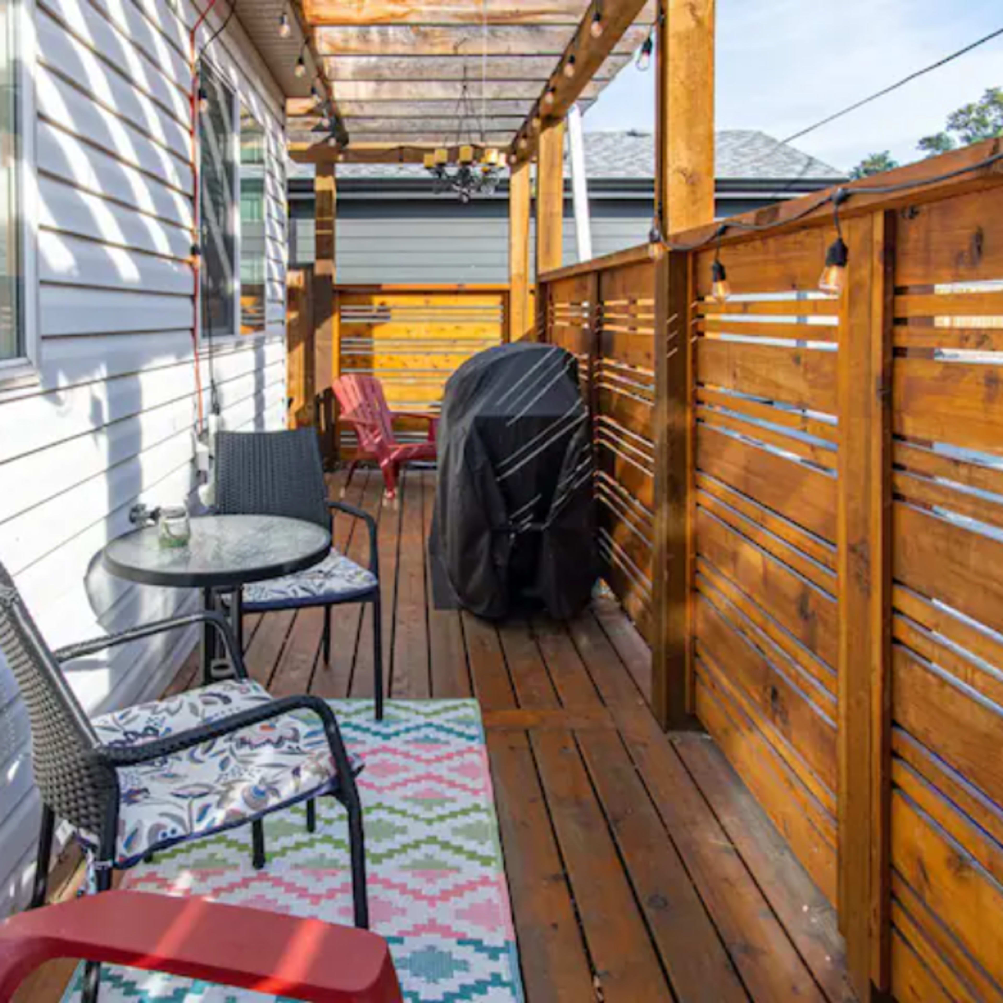 A wooden deck with two chairs, a small table, and a barbecue grill, surrounded by a wood railing under a pergola.