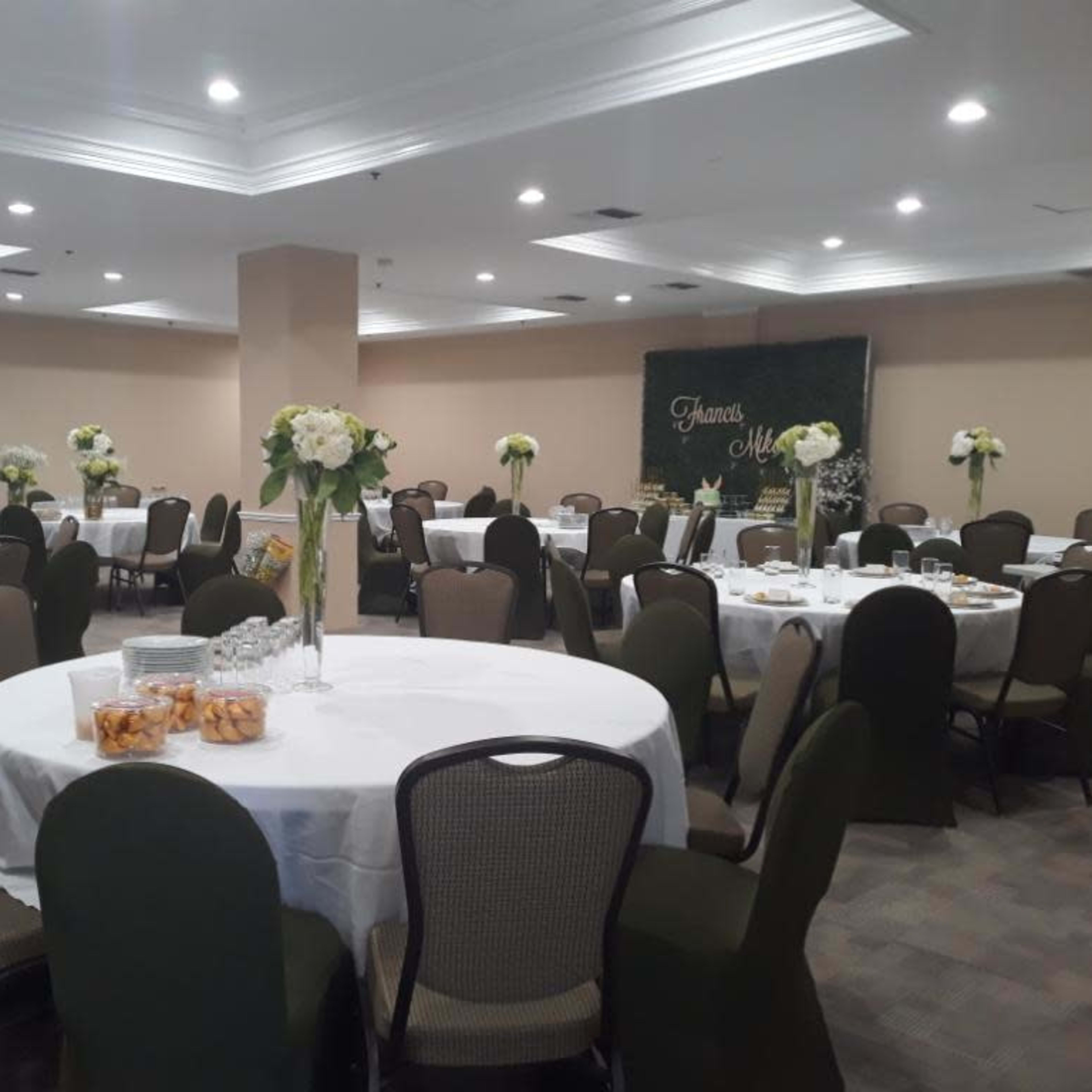 A banquet room is set up with round tables covered in white tablecloths and topped with floral centerpieces, surrounded by green chairs.
