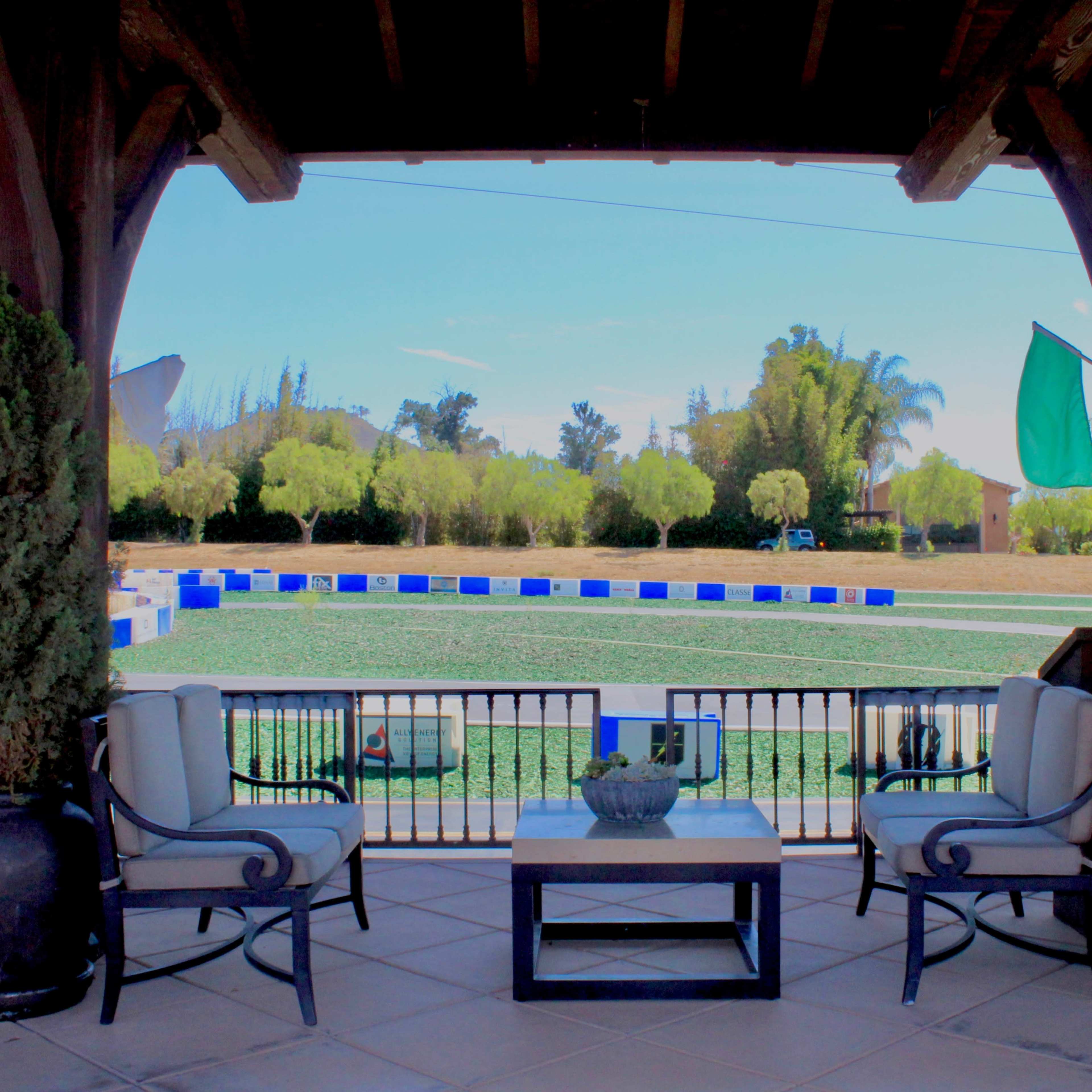 A porch with two chairs and a table overlooking a green field surrounded by blue and white barriers.
