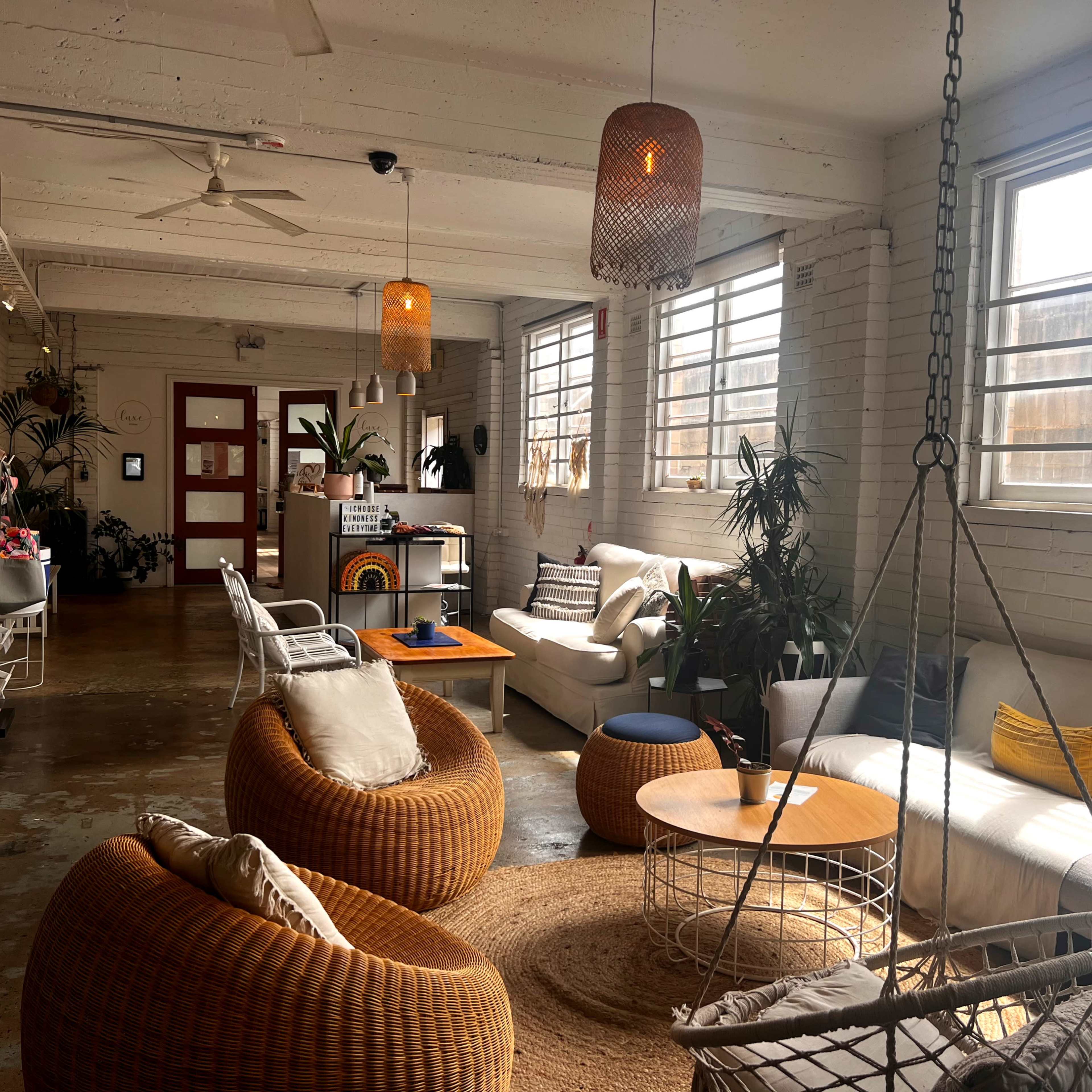 A spacious, well-lit lounge area featuring a variety of seating options, including woven chairs and sofas, along with potted plants and large windows.