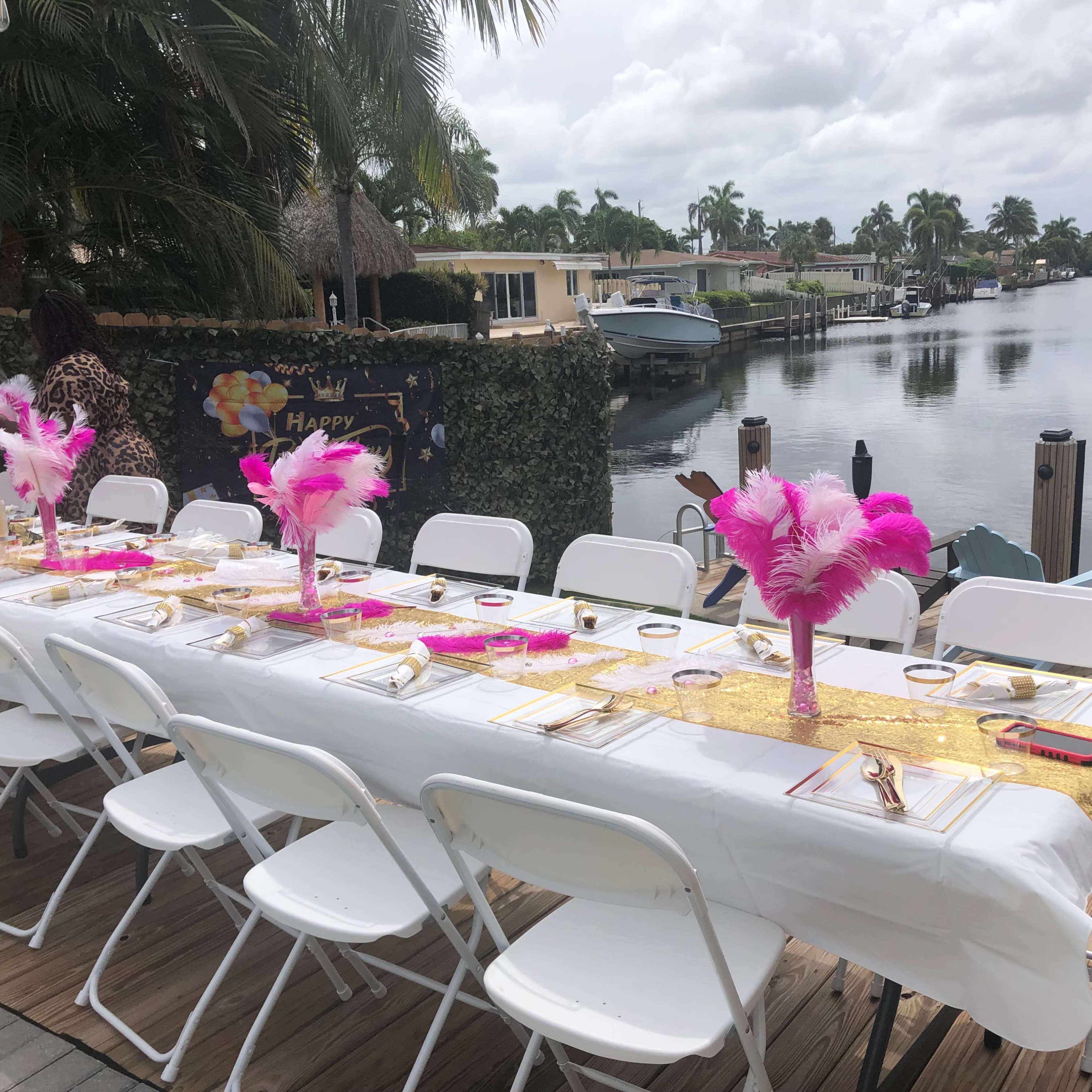 A long, decorated dining table with pink feather centerpieces is set up by a waterfront lined with boats and palm trees.