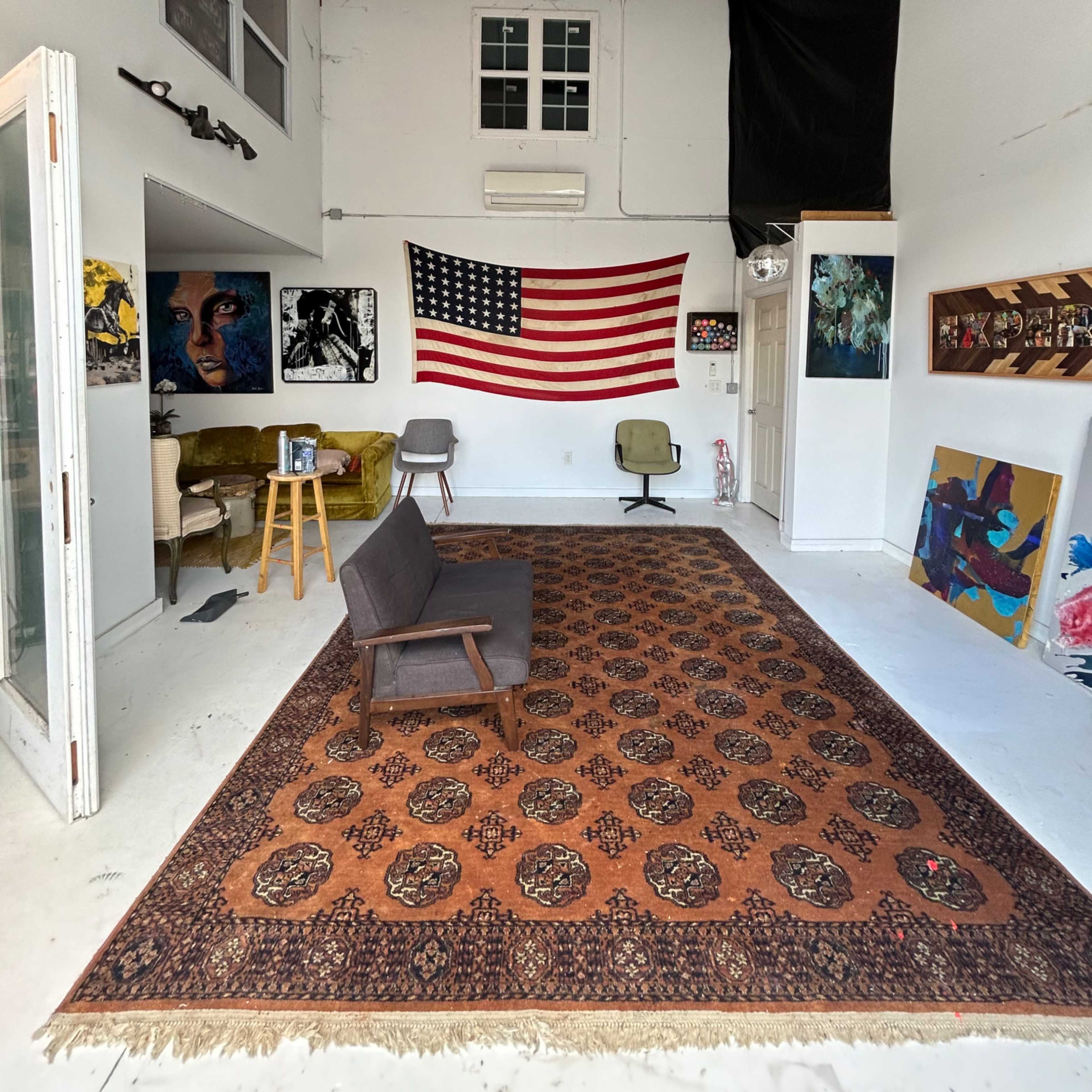 An art studio with a large rug, a U.S. flag on the wall, various artworks, and a few pieces of furniture, including chairs and a small table.