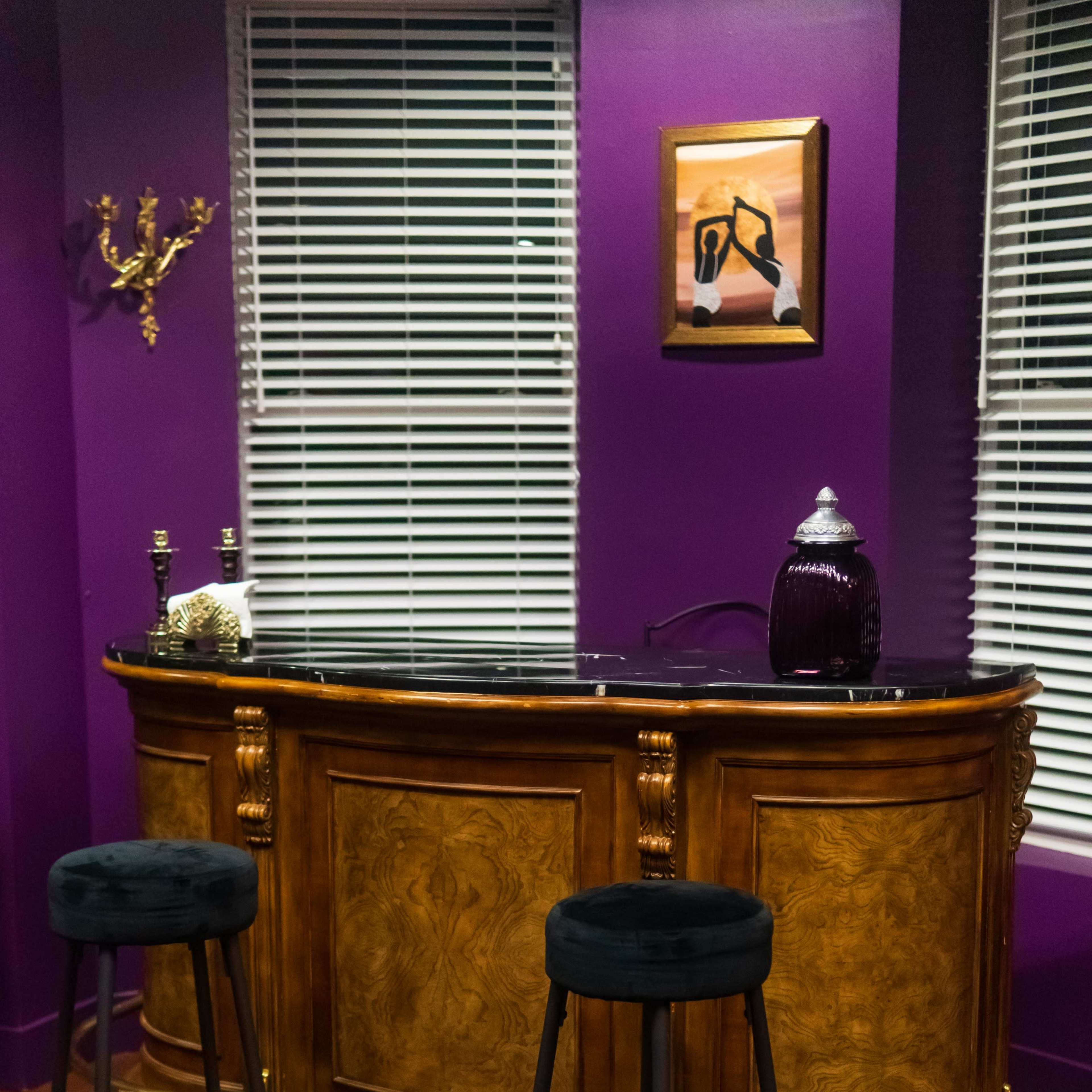 A wooden bar with a black countertop and two black stools in a room with purple walls and white blinds.