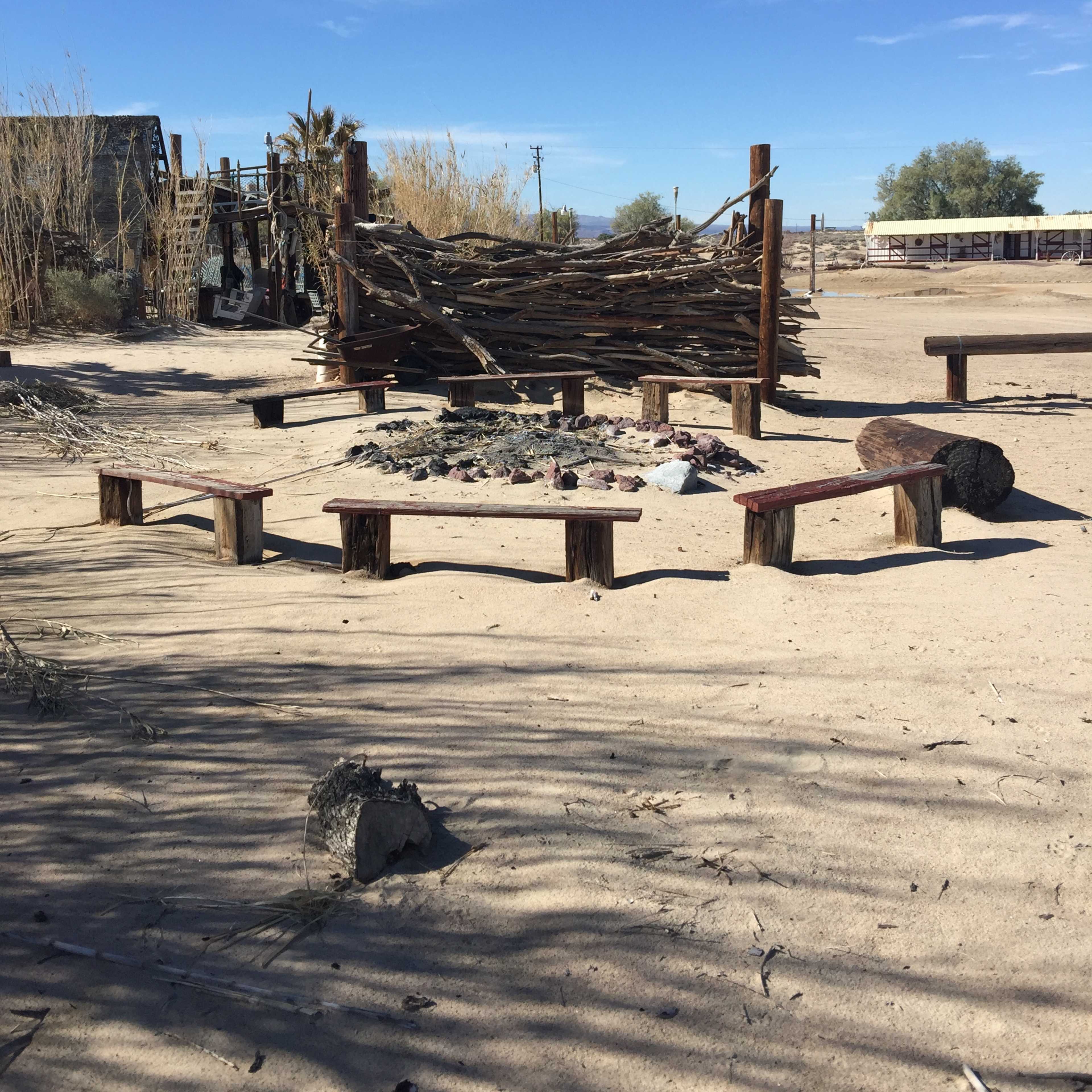 A sandy area features a circular seating arrangement made of logs surrounding a stone fire pit, with a wooden structure in the background.