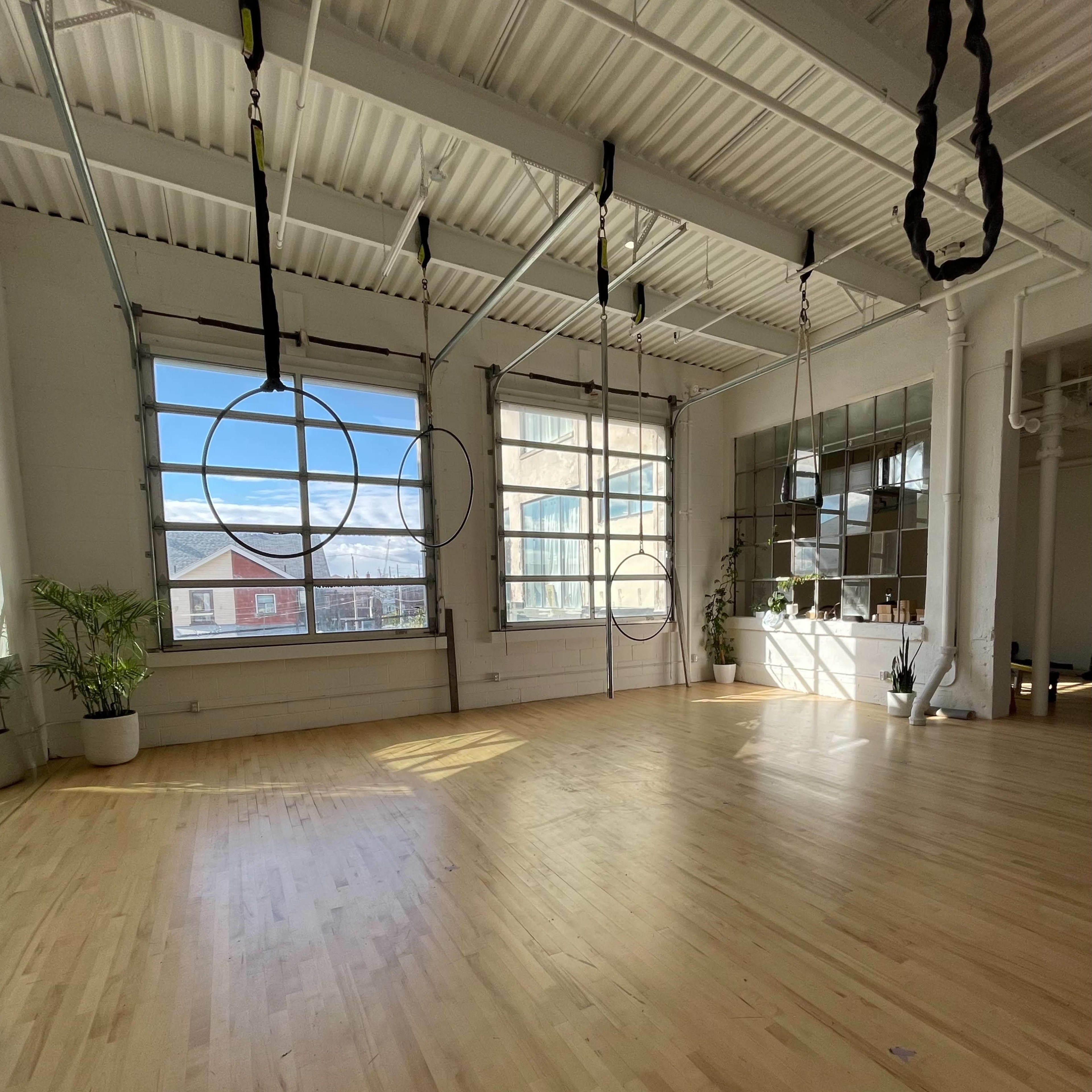 A spacious, well-lit studio with wooden flooring and aerial hoops suspended from the ceiling.