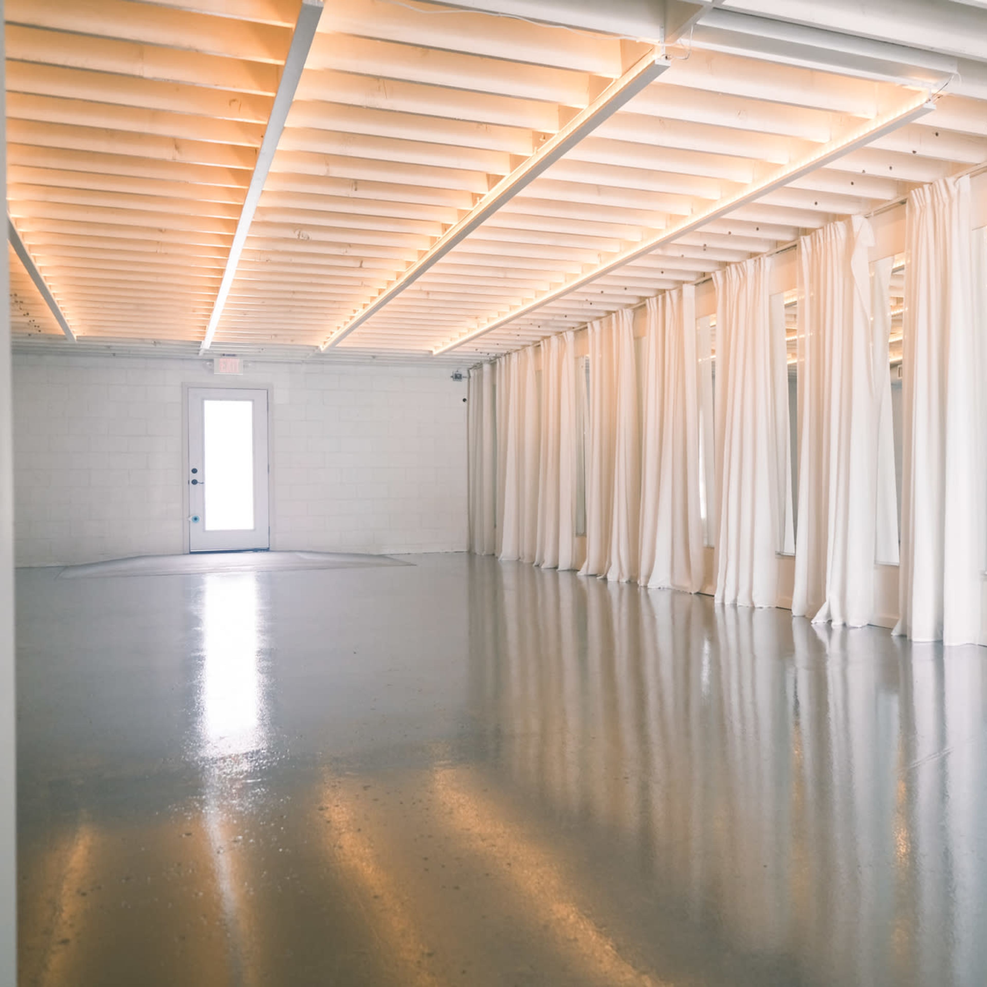 A spacious, empty room with a polished floor, white curtains along the walls, and natural light coming through a door at one end.