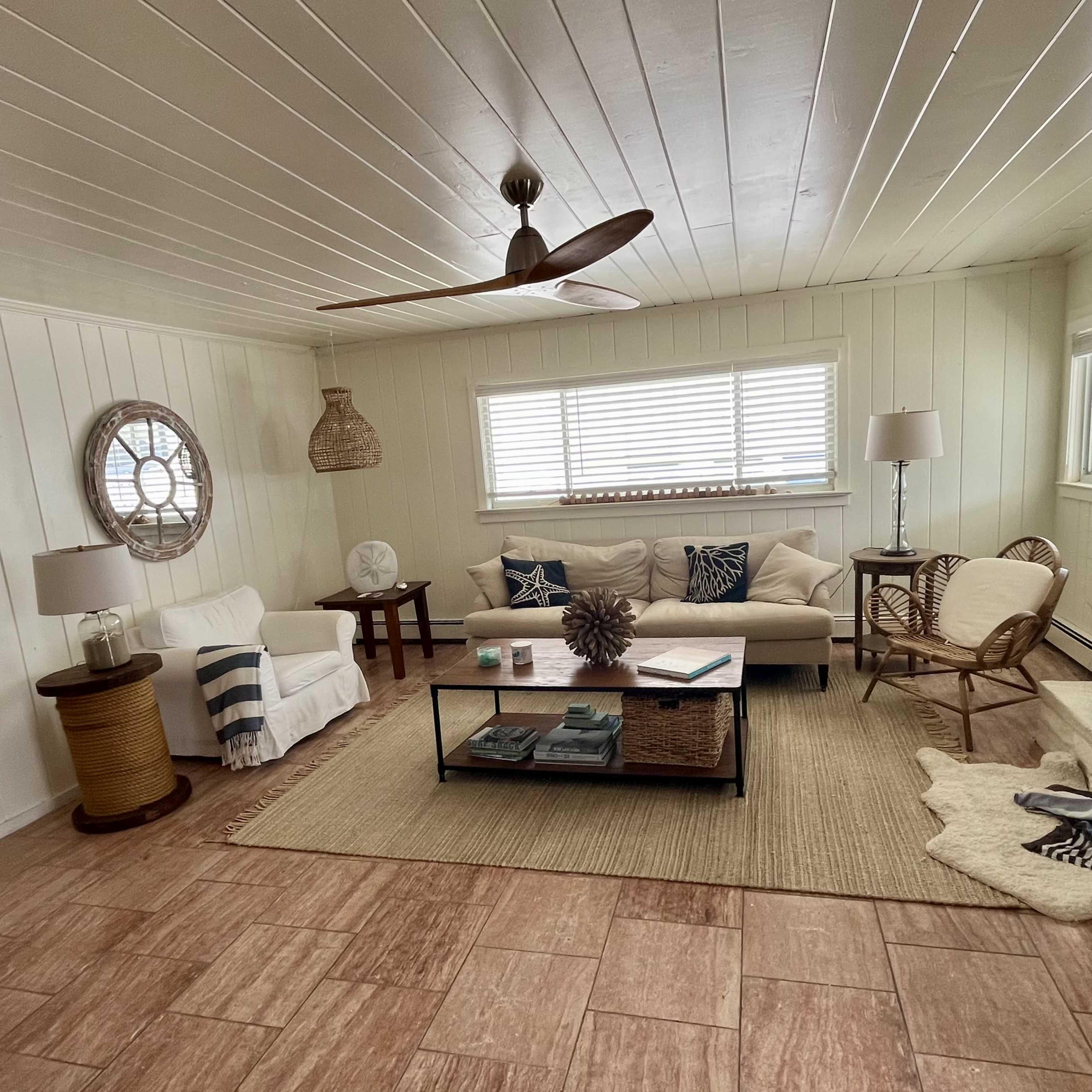 The living room features a light-colored sofa, a coffee table with books, a woven rug, and a fireplace surrounded by natural light from several windows.