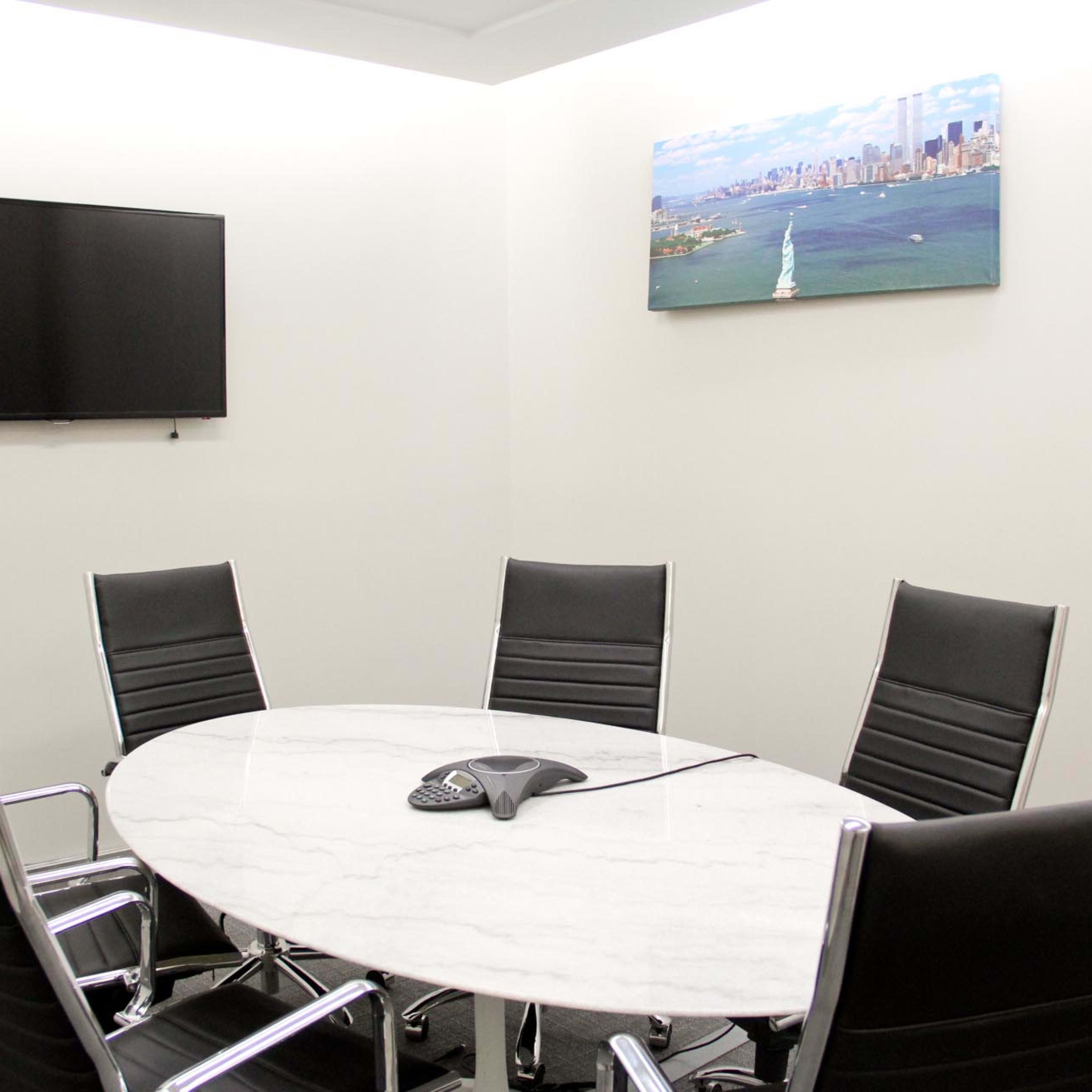 A modern conference room features a round marble table surrounded by six black chairs, with a television and a large image of a cityscape on the wall.
