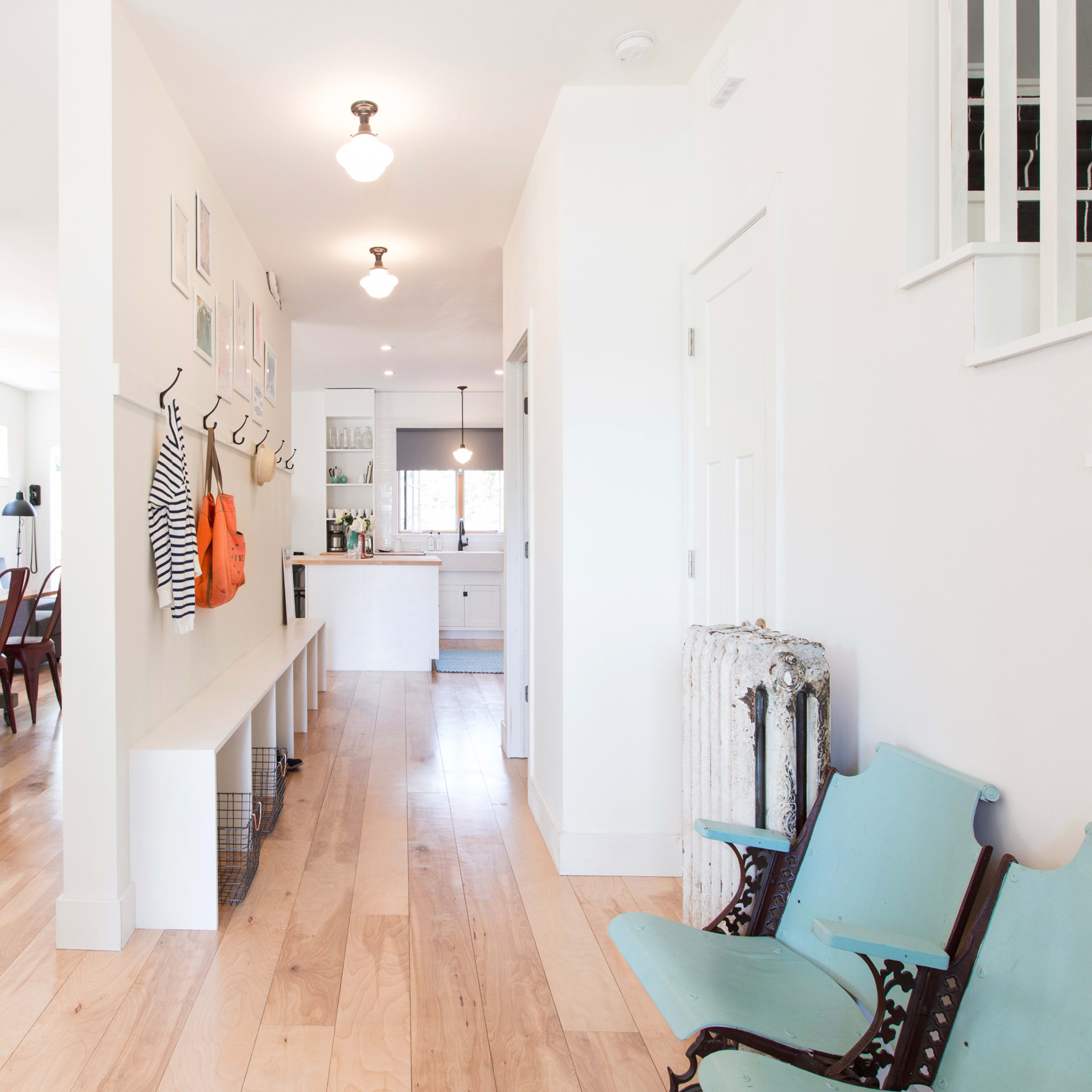 A bright, open hallway leading to a modern kitchen and dining area, with wooden floors and a seating area featuring vintage-looking chairs.