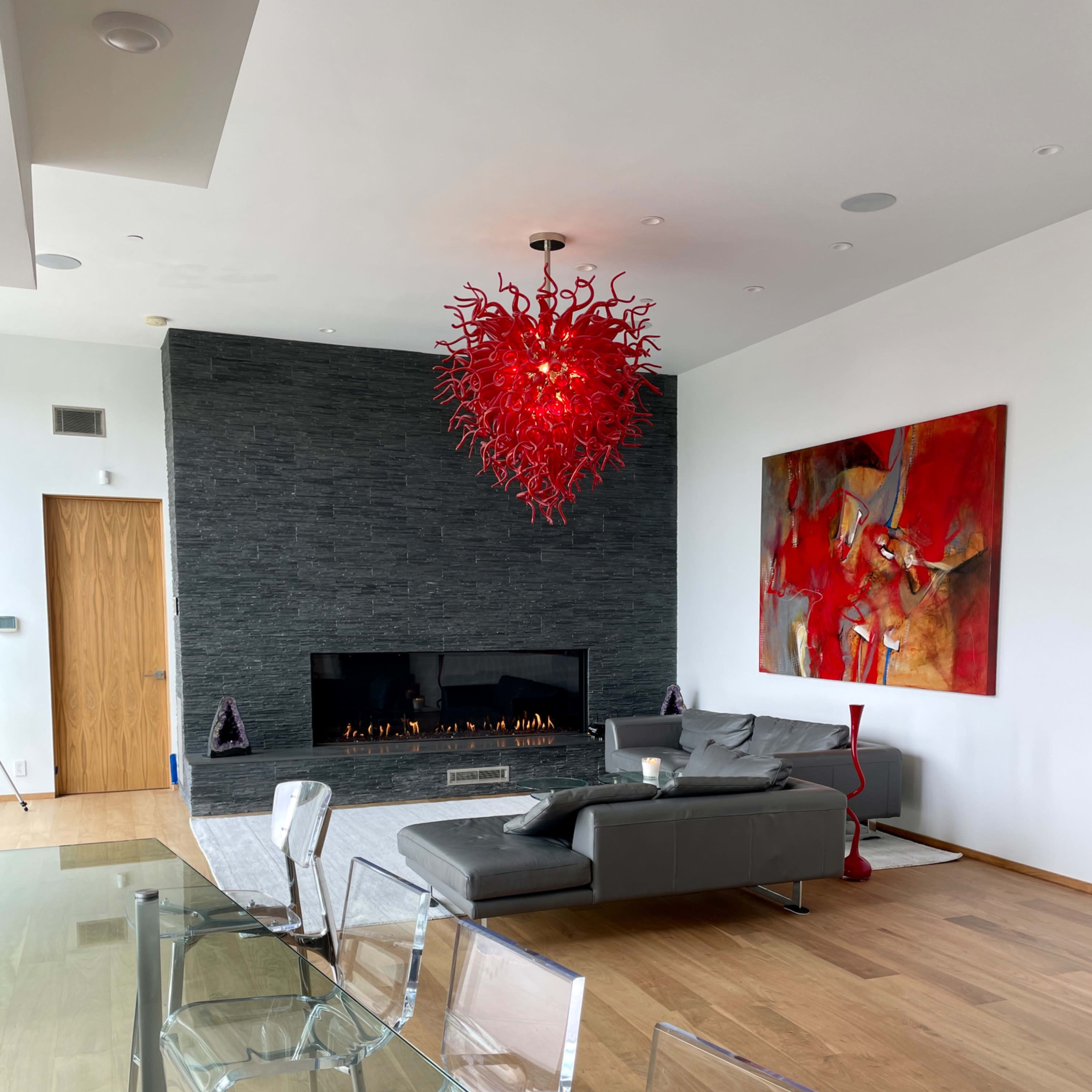 A modern living room featuring a stone fireplace, a large red chandelier, and minimalist furniture, along with an abstract painting on the wall.