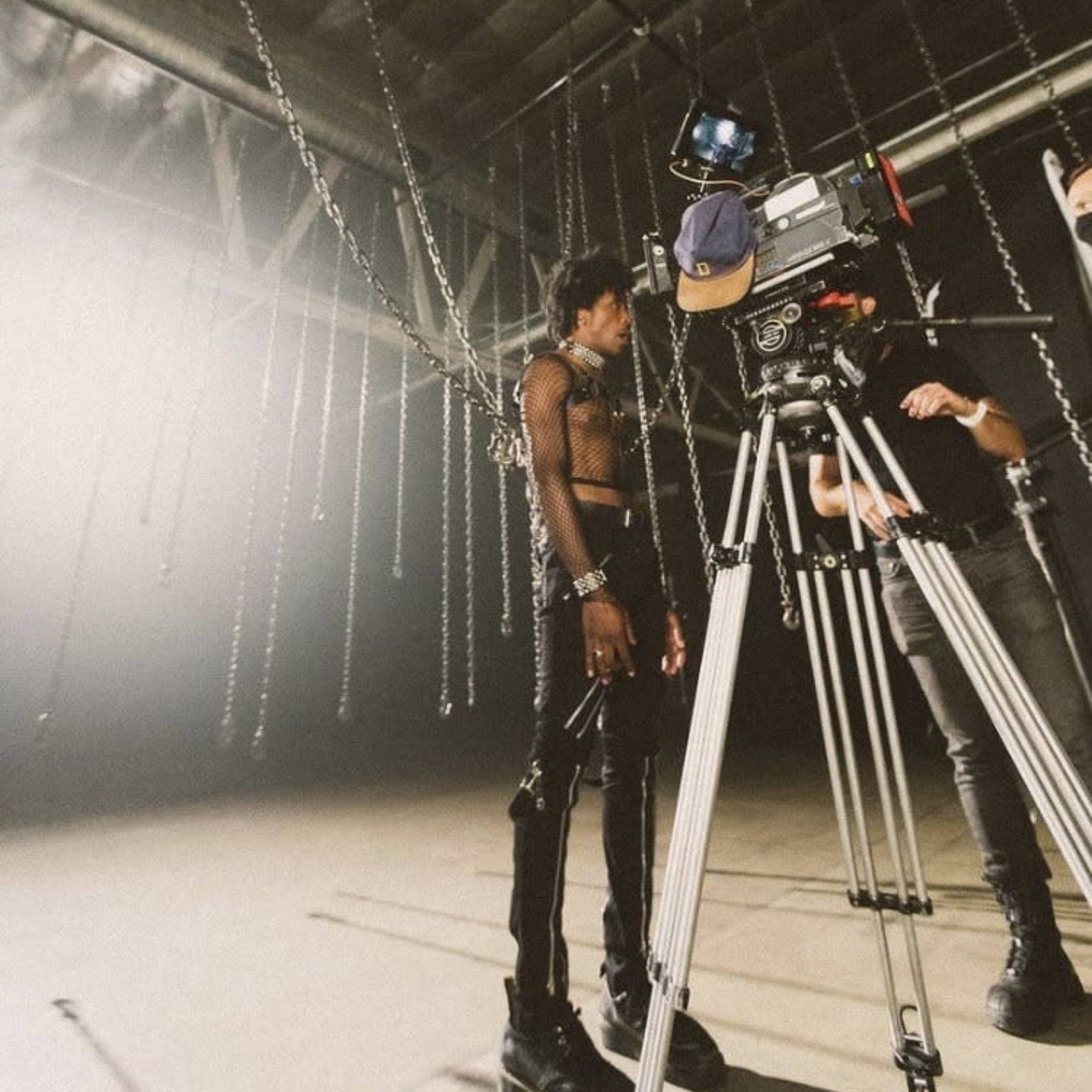 A person in a mesh outfit stands near a camera on a tripod in a dimly lit studio with chains hanging from the ceiling.