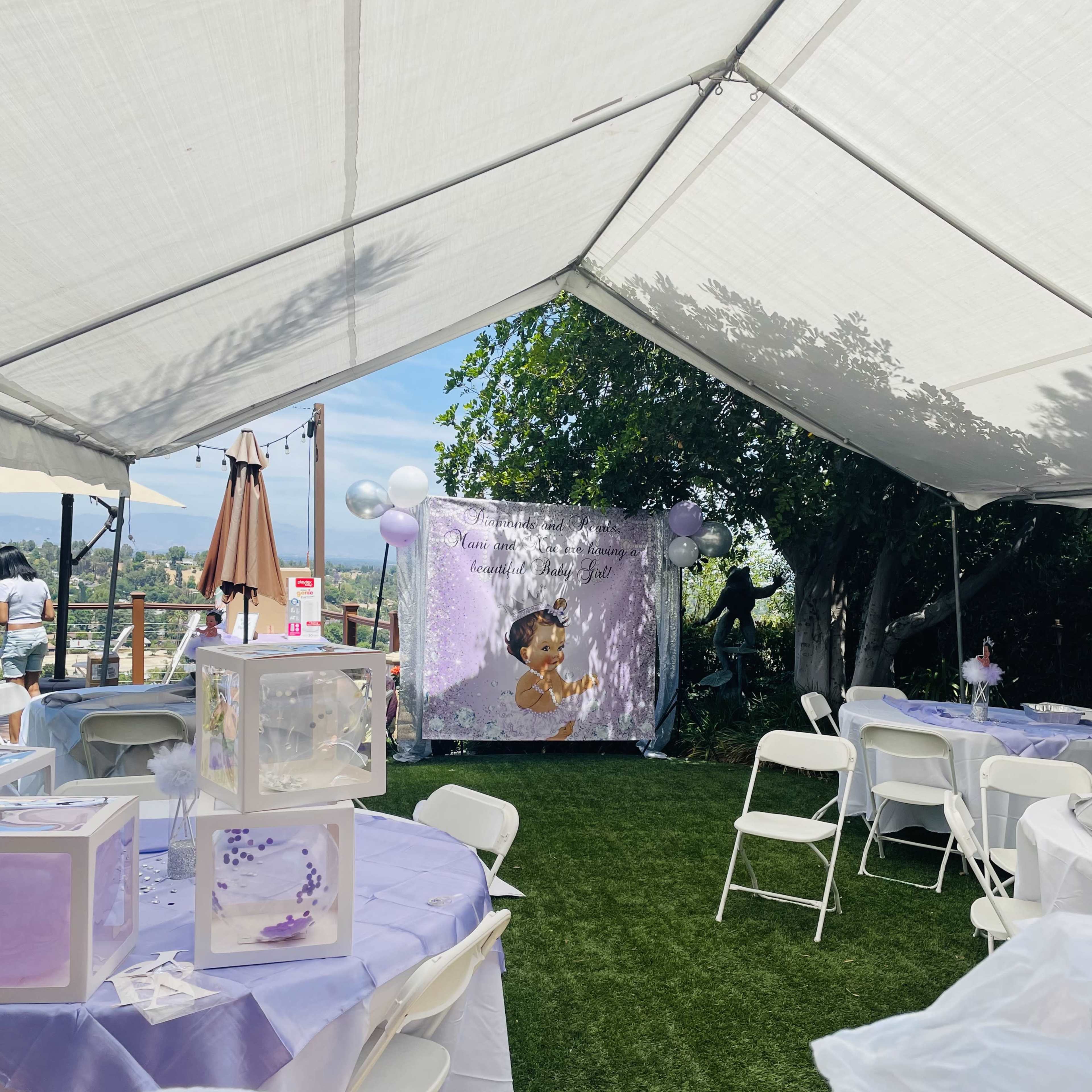 A decorated outdoor space features tables covered in lavender cloth and a backdrop with a child's image, surrounded by balloons and greenery.
