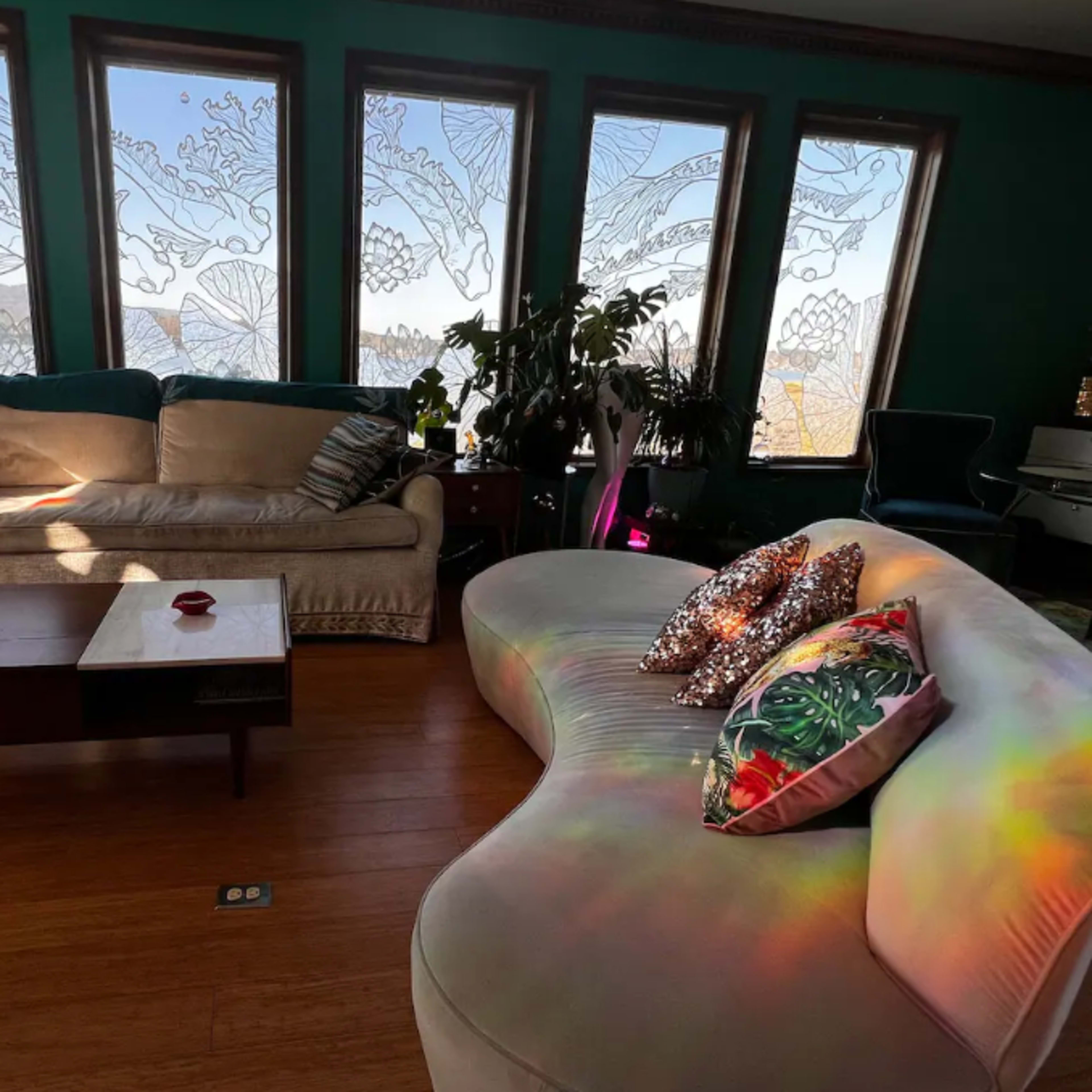 A living room with two distinct sofas, large windows adorned with intricate designs, and various plants scattered throughout the space.