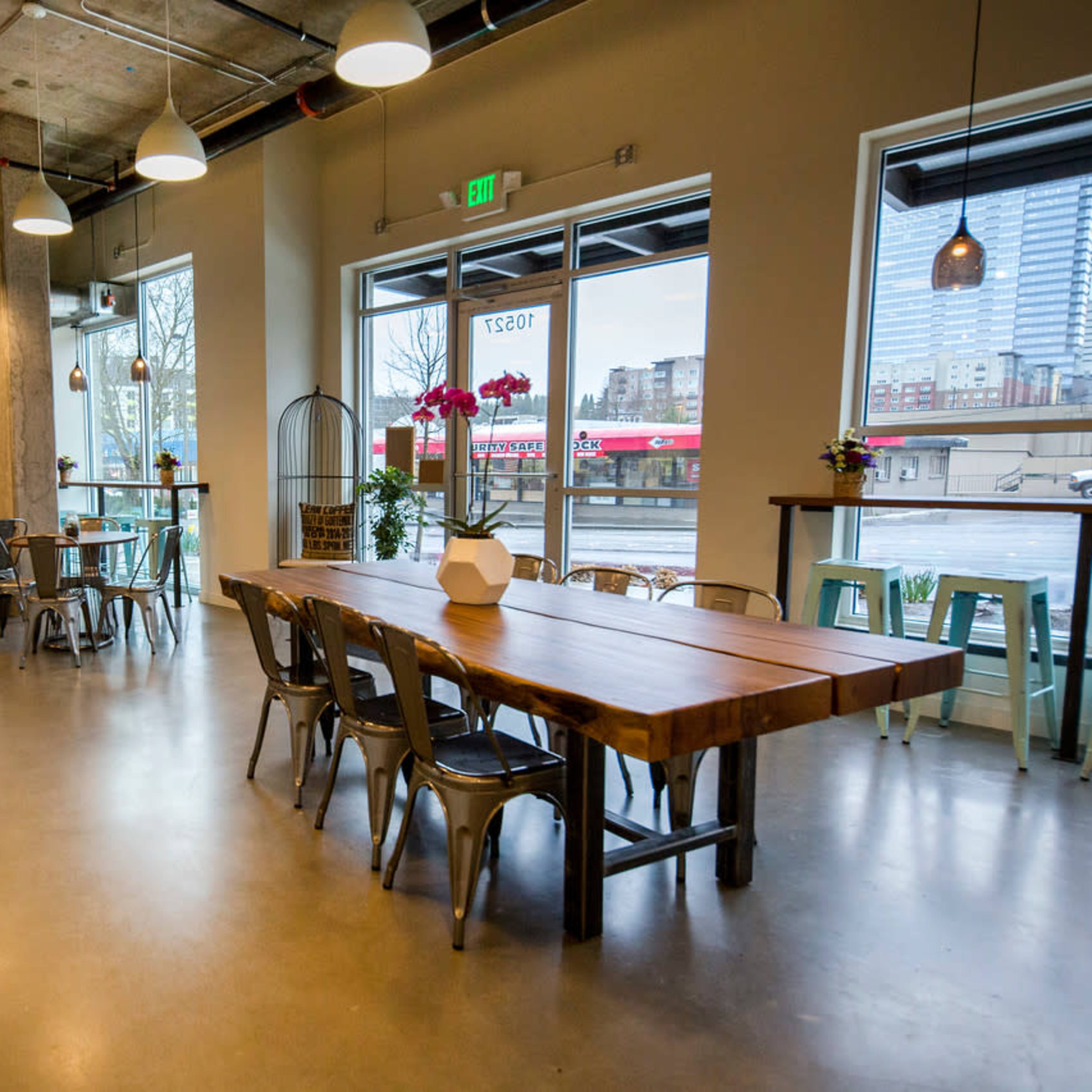 A modern café interior with a long wooden table, metal chairs, and large windows offering a view of the street outside.