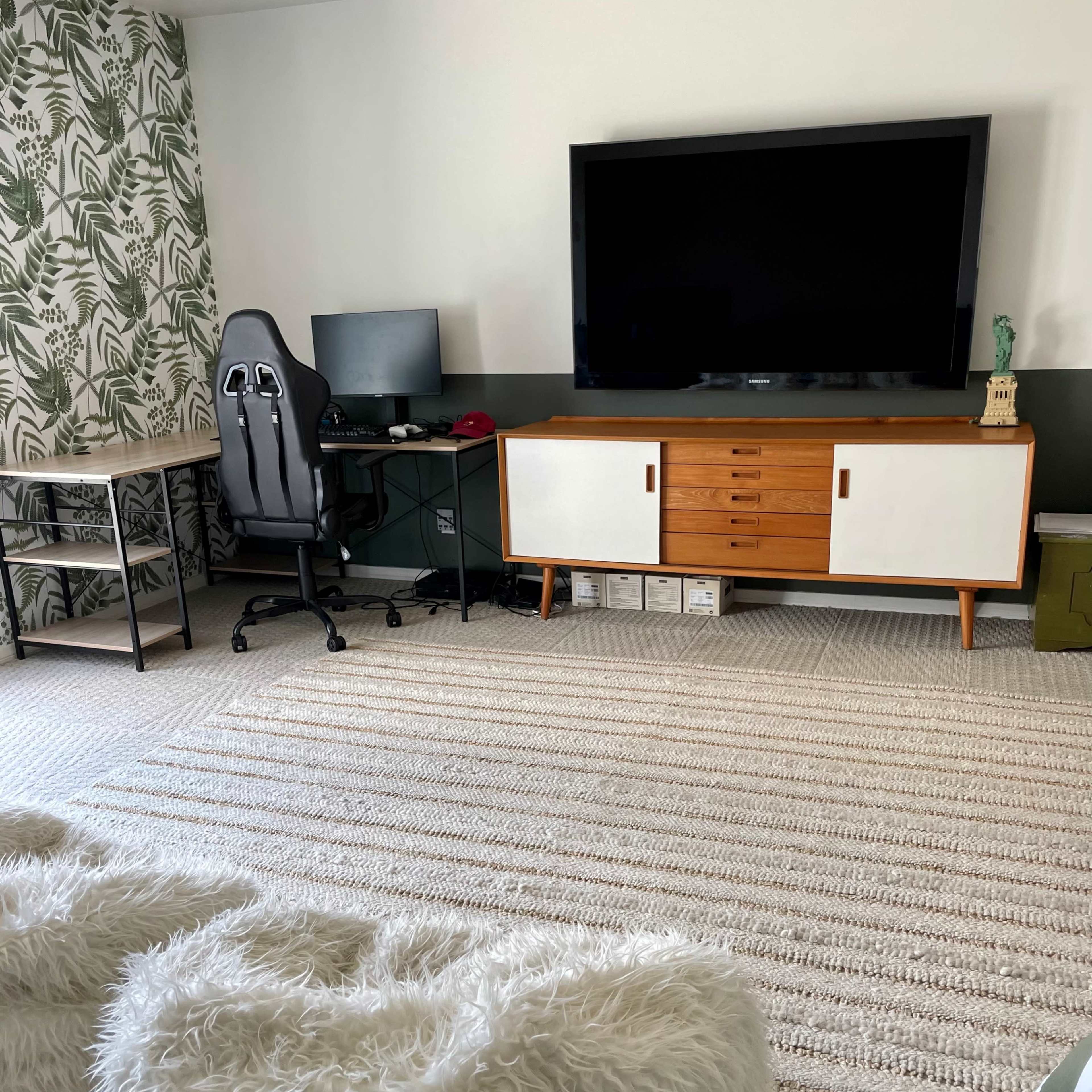 A living space with a television mounted on the wall, a wooden media cabinet, a desk with a computer, an office chair, and two fluffy white poufs on a textured rug.