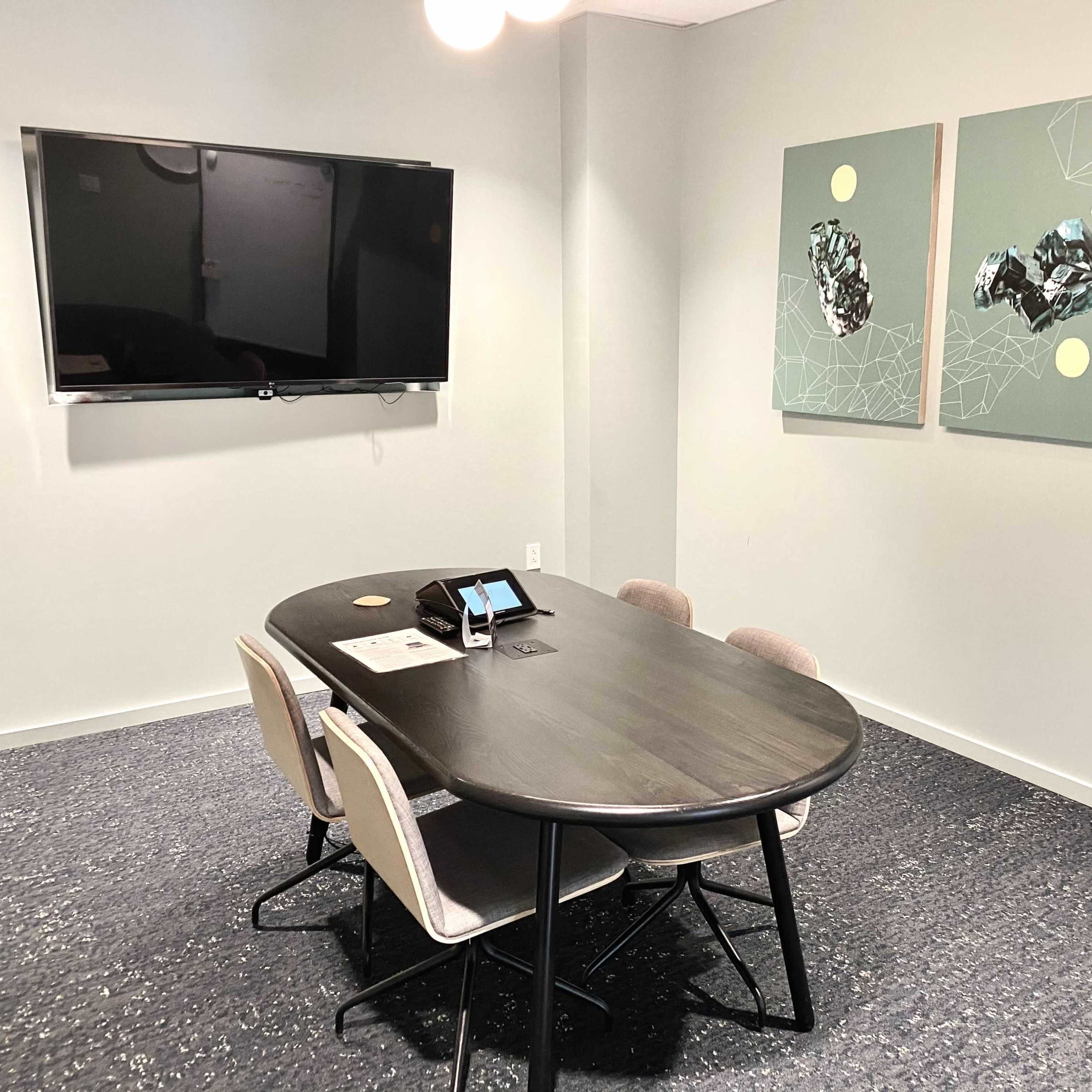 A modern meeting room featuring a round table with four chairs, a large wall-mounted television, and three abstract art pieces hanging on the wall.