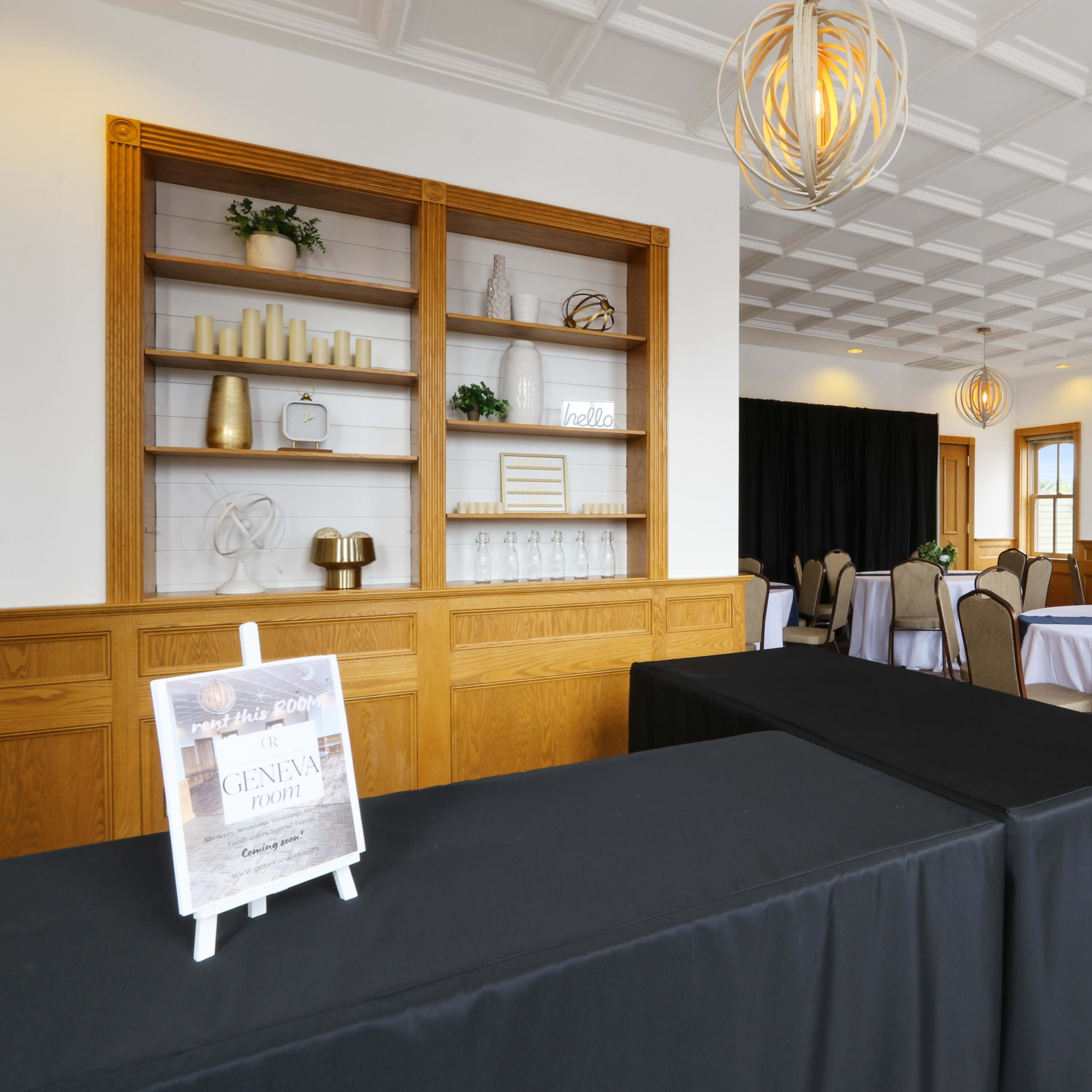 A well-lit event space featuring tables with beige chairs, a black table in the foreground, and a decorative shelving unit against the wall.