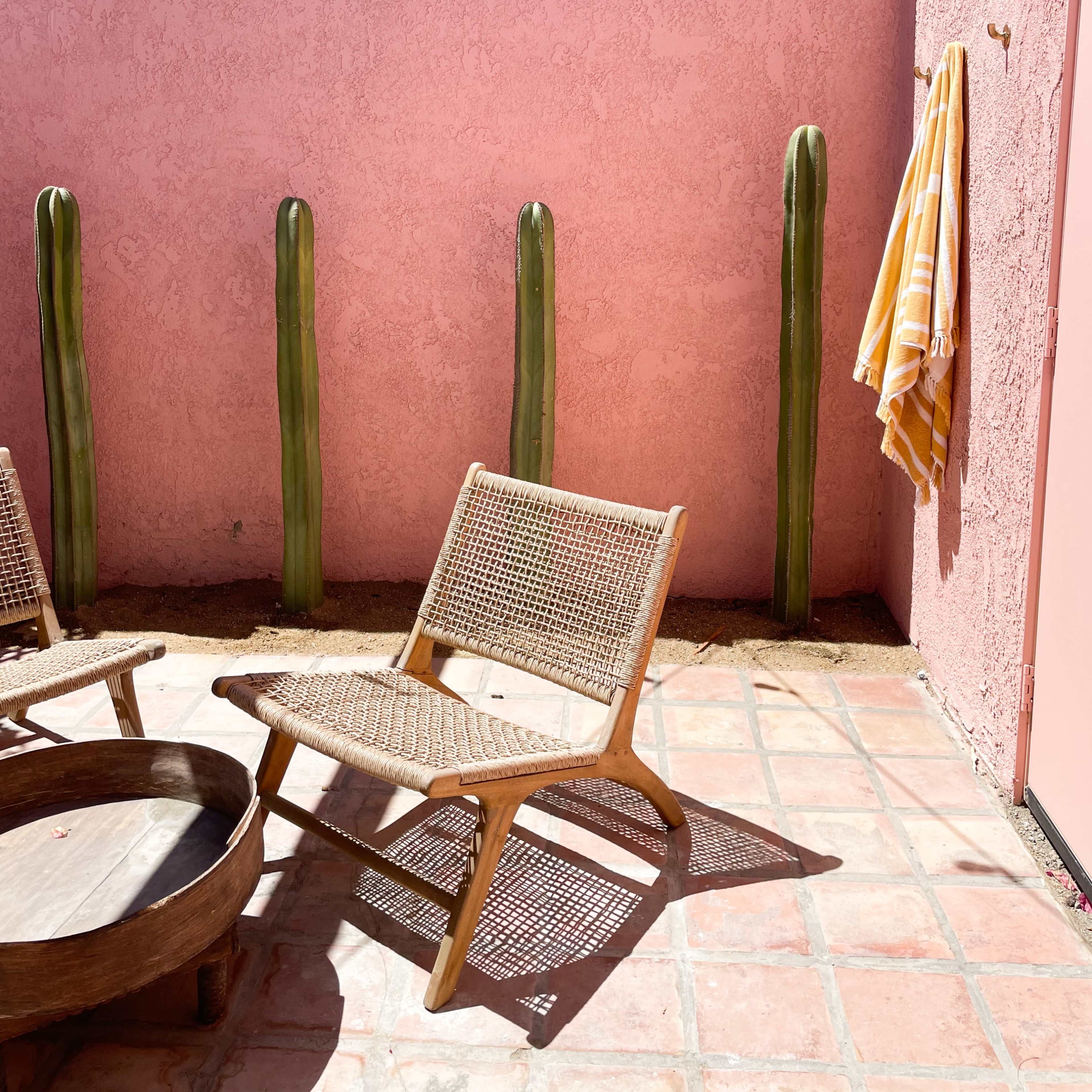 A small outdoor patio features a pink wall, two wooden chairs, a circular table, and tall cacti along one side.