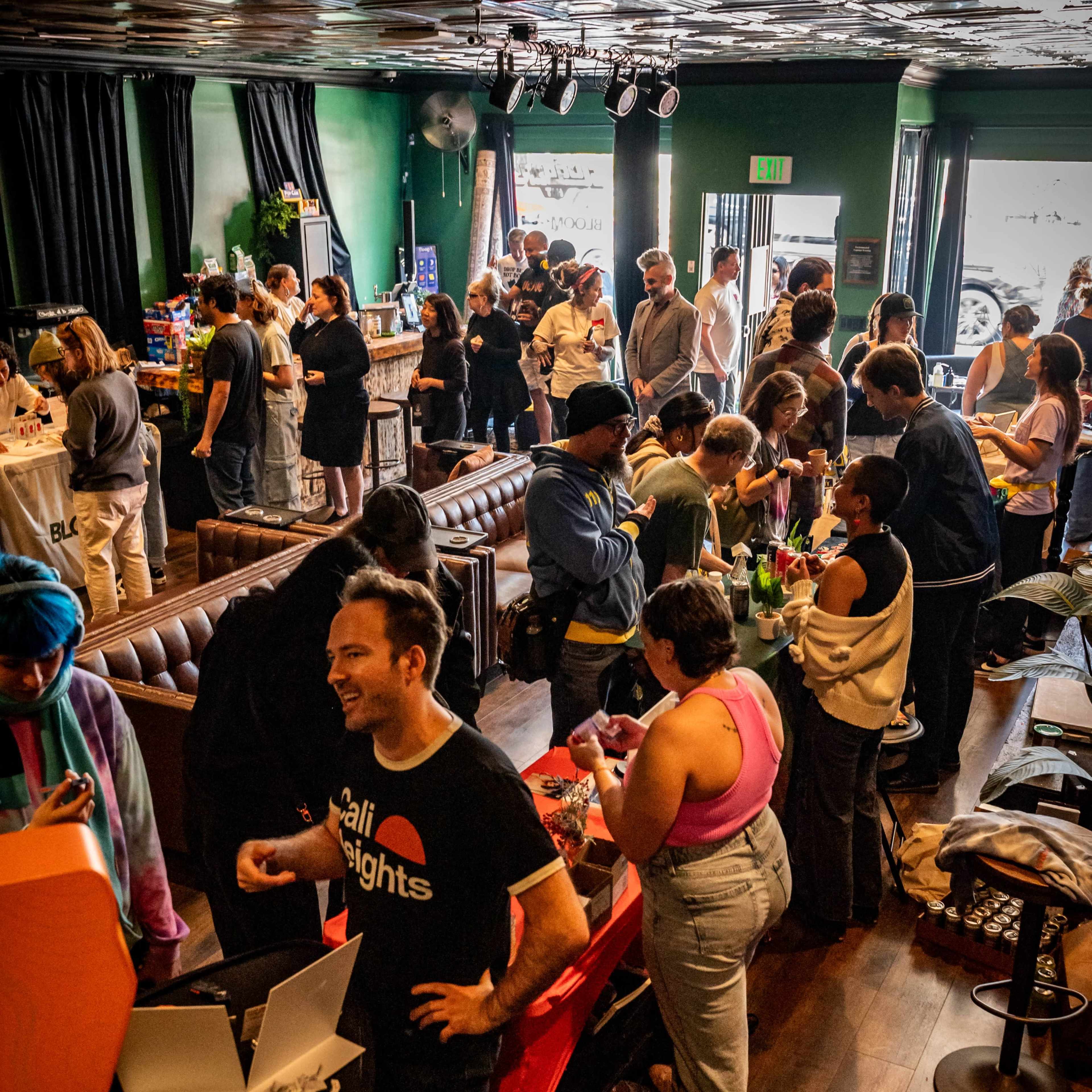 A large group of people mingles in a lively venue filled with tables displaying food and drinks, while others engage in conversation.
