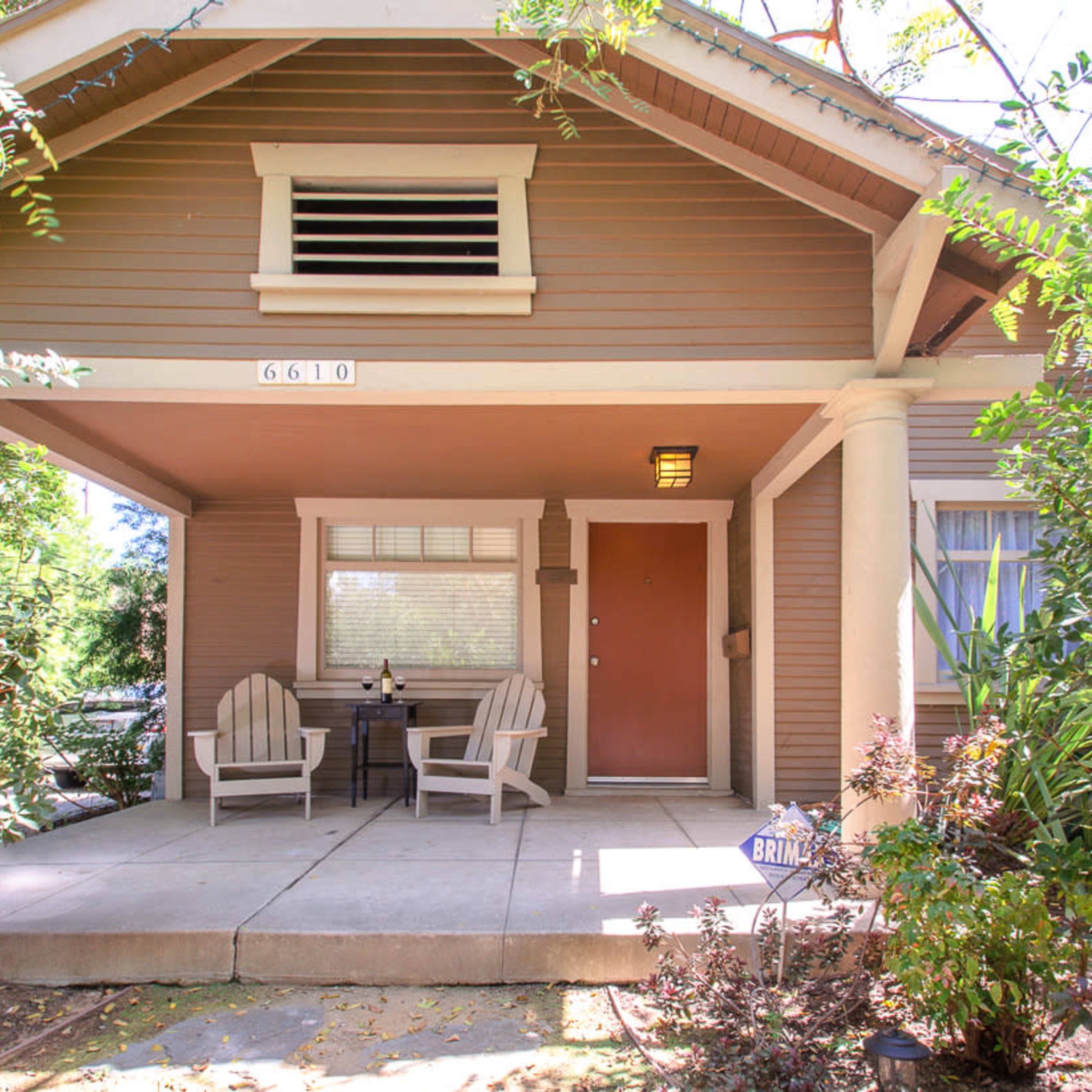 A front porch of a house with two chairs and a small table, surrounded by greenery.