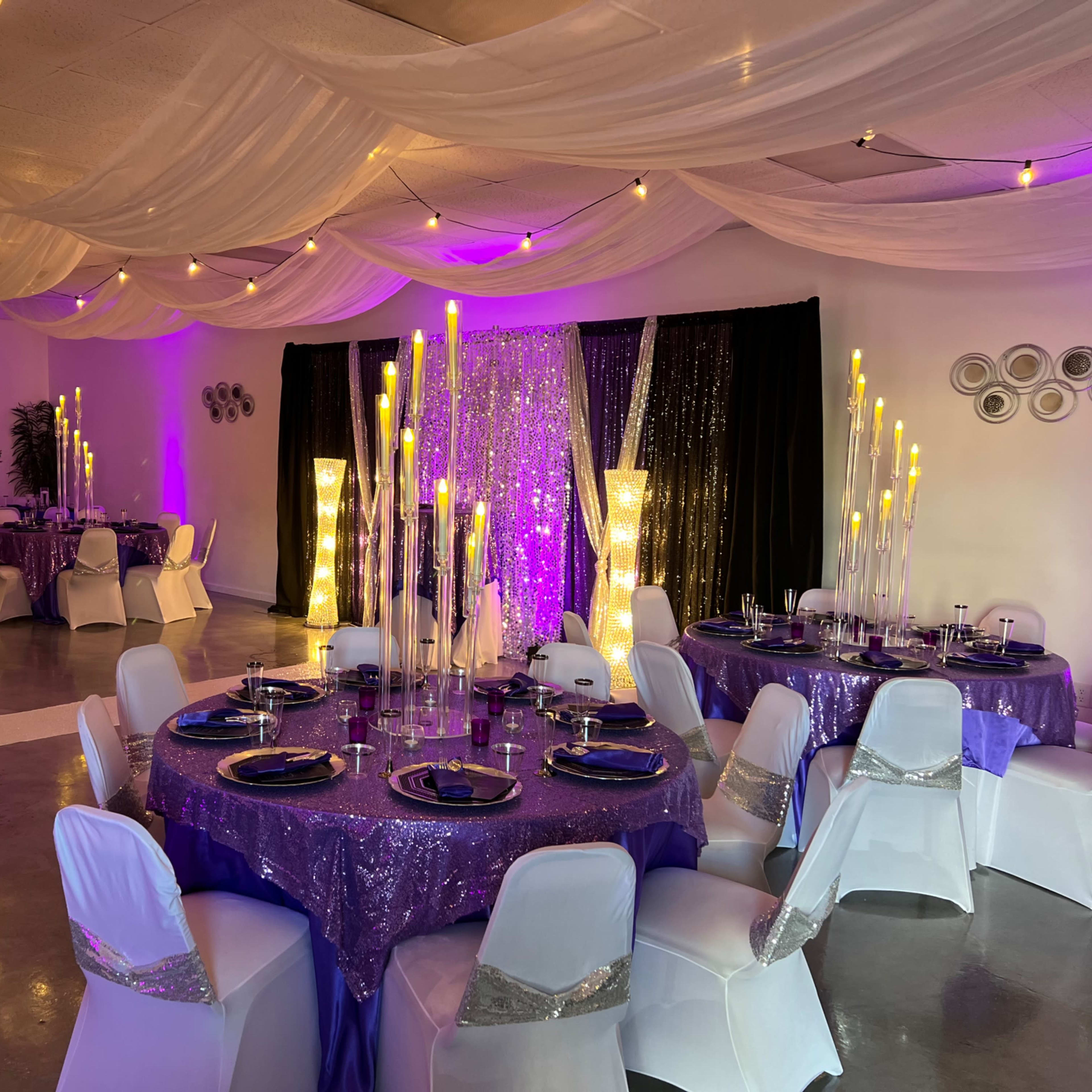 A decorated event space with tables covered in purple cloth, elegant candle centers, and ambient lighting from draped fabric and string lights.