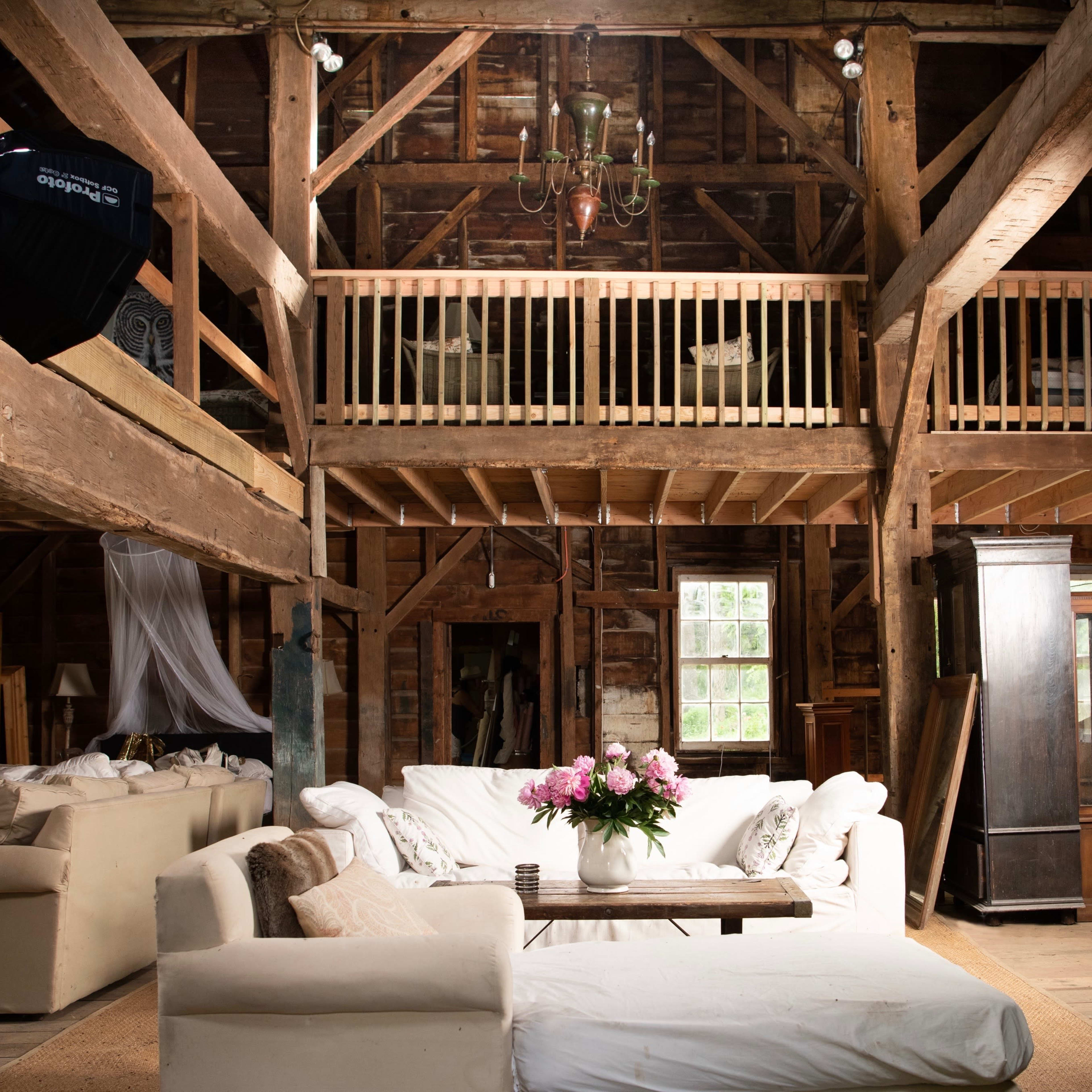 A spacious, rustic barn interior features a seating area with white sofas and a flower arrangement on a wooden coffee table, complemented by a large softbox light and a lofted section above.