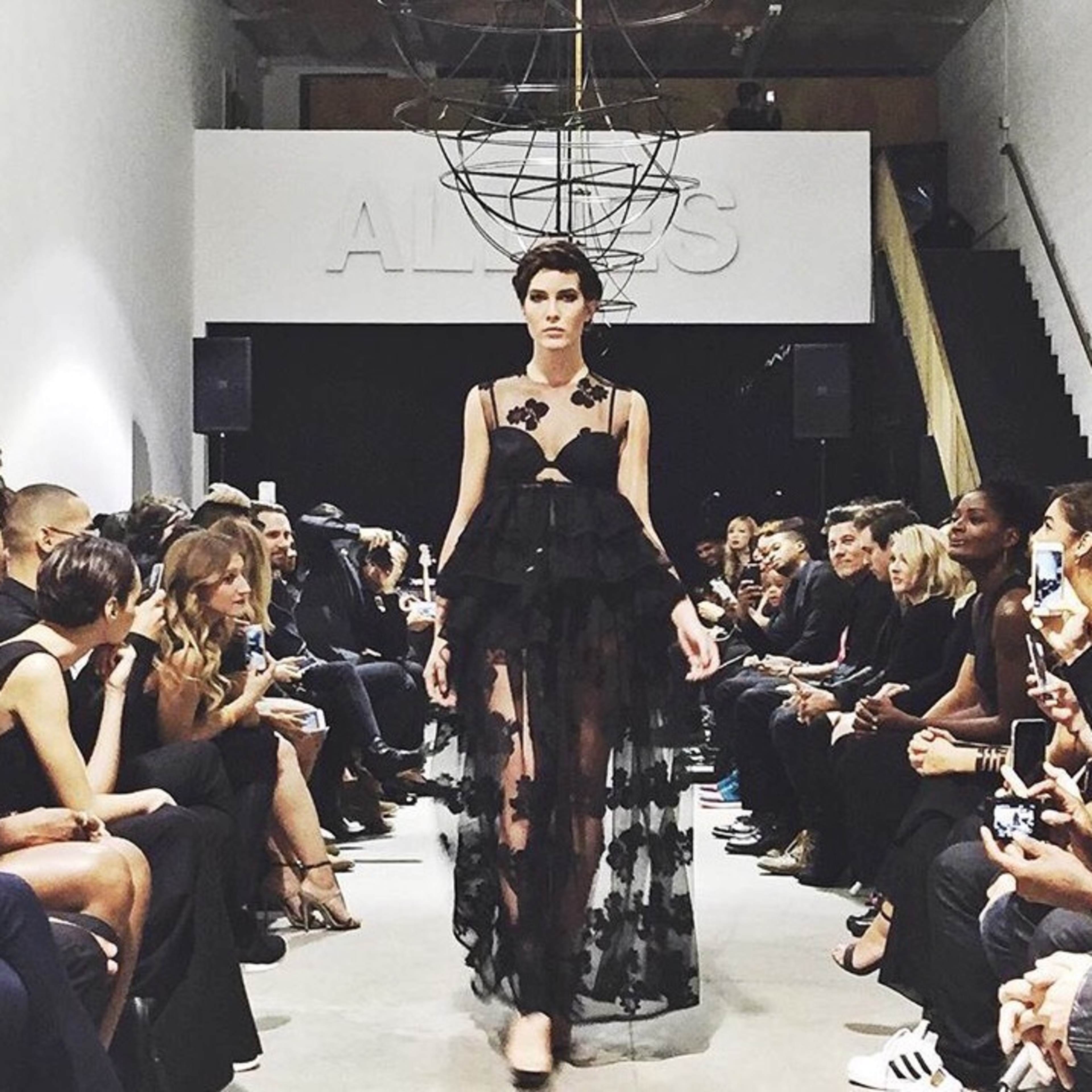 A model walks down a runway in a fashion show while an audience watches from both sides.