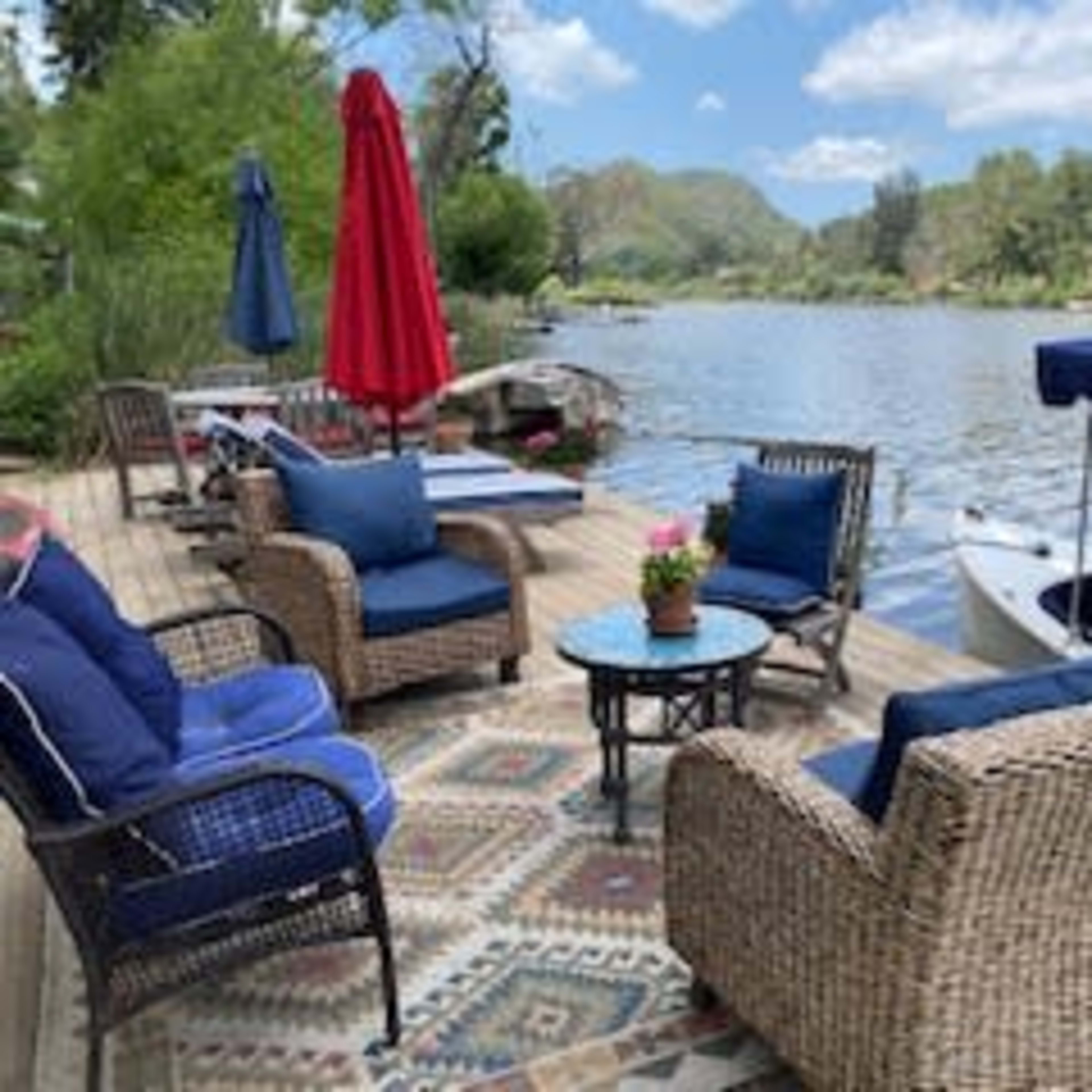 A cozy seating area with wicker chairs and a small table overlooks a tranquil river surrounded by hills.