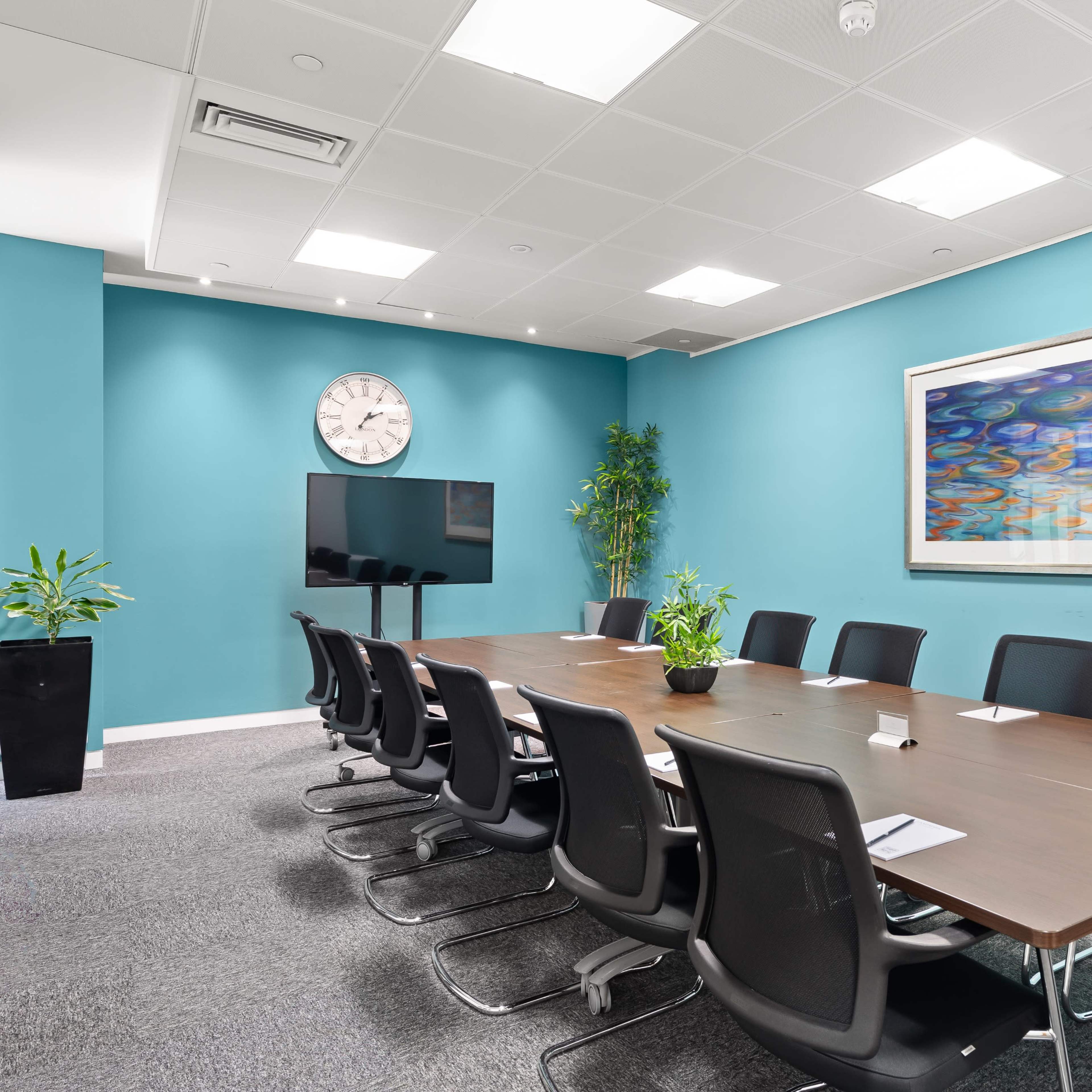 A modern conference room features a long wooden table surrounded by black chairs, a large television, a wall clock, and colorful artwork, all set against a vibrant blue wall.