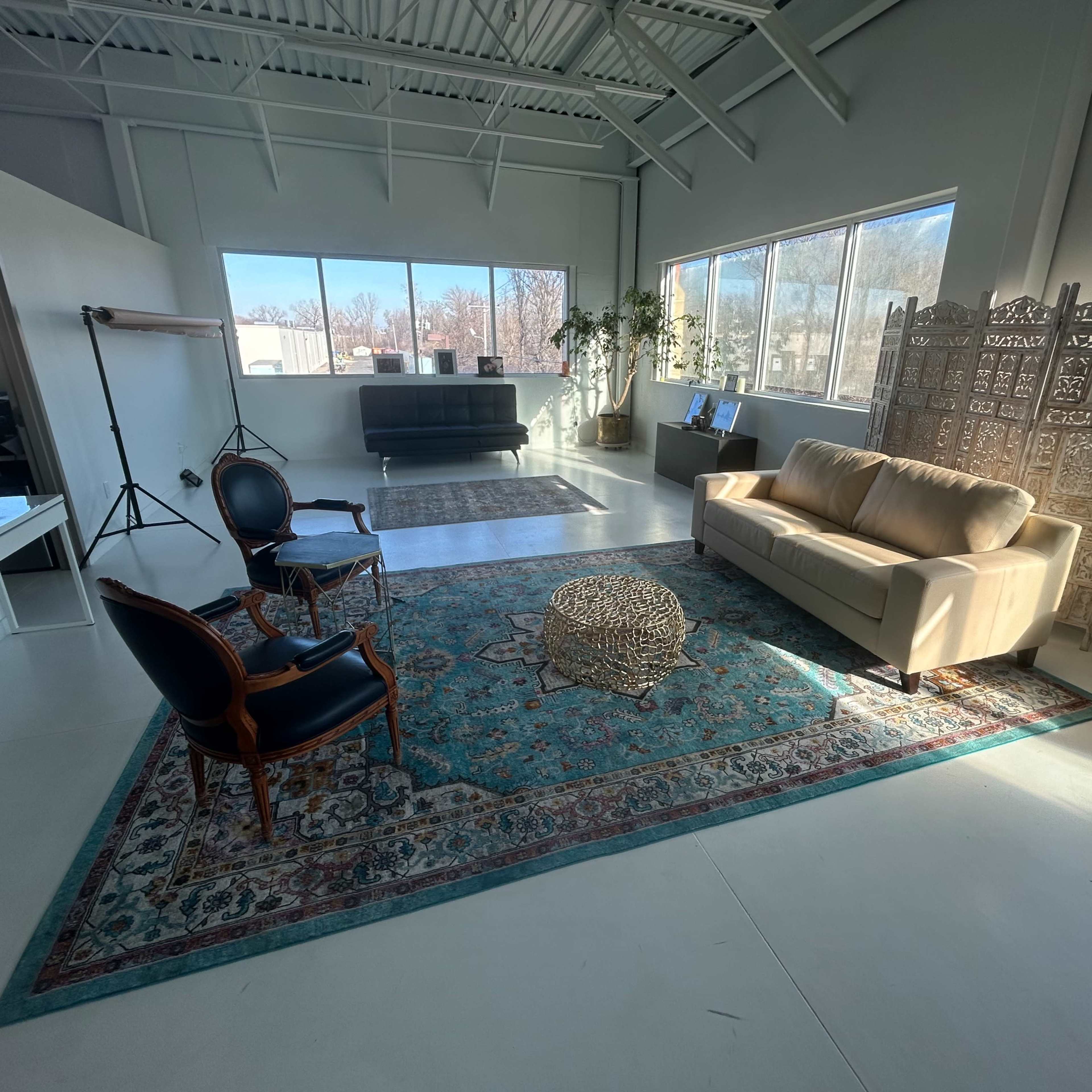 A spacious, well-lit room featuring a beige sofa, a decorative rug, two chairs, and a partition, with large windows allowing natural light to enter.