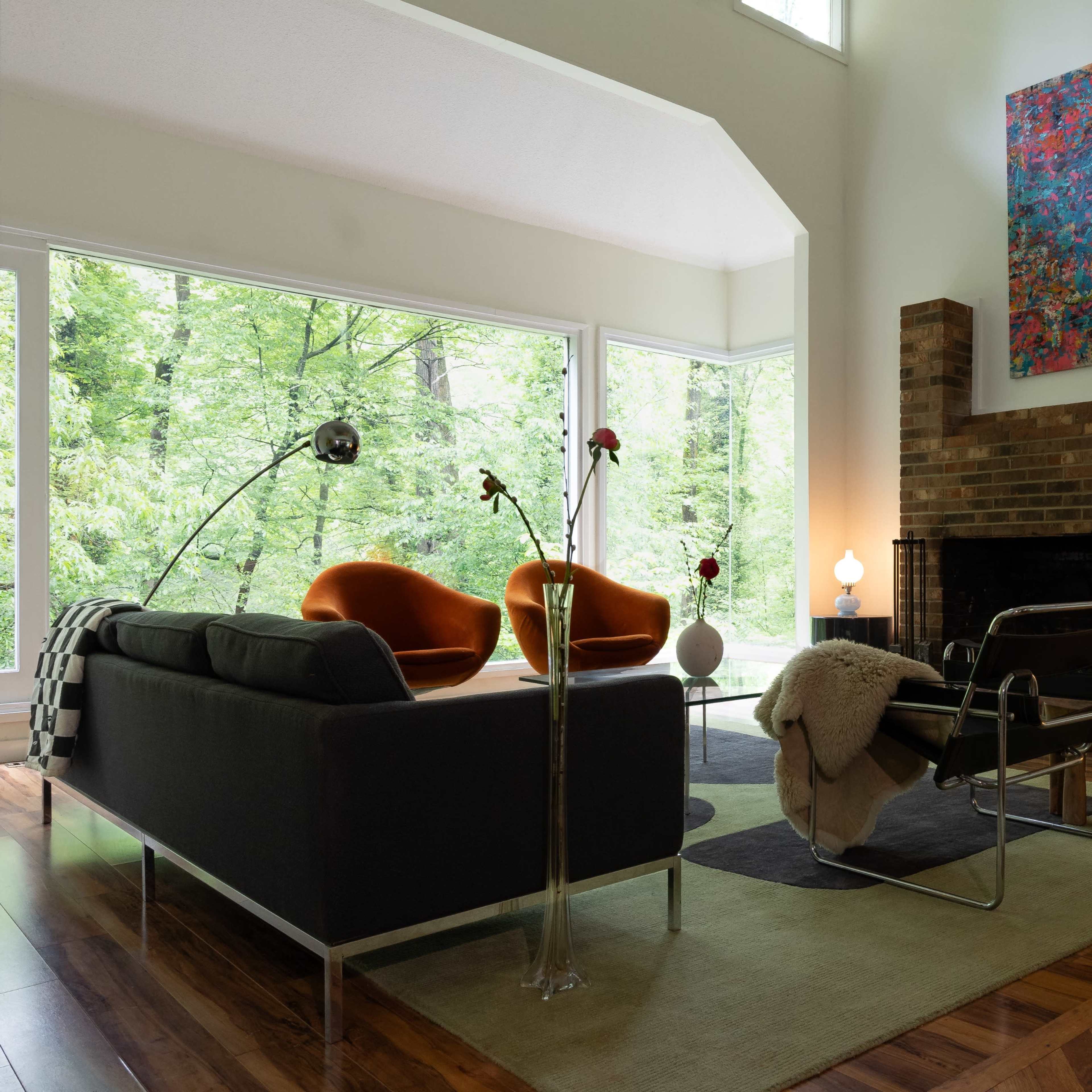 A modern living room features large windows with a view of greenery, two orange chairs, a black couch, and a brick fireplace with a colorful artwork above it.