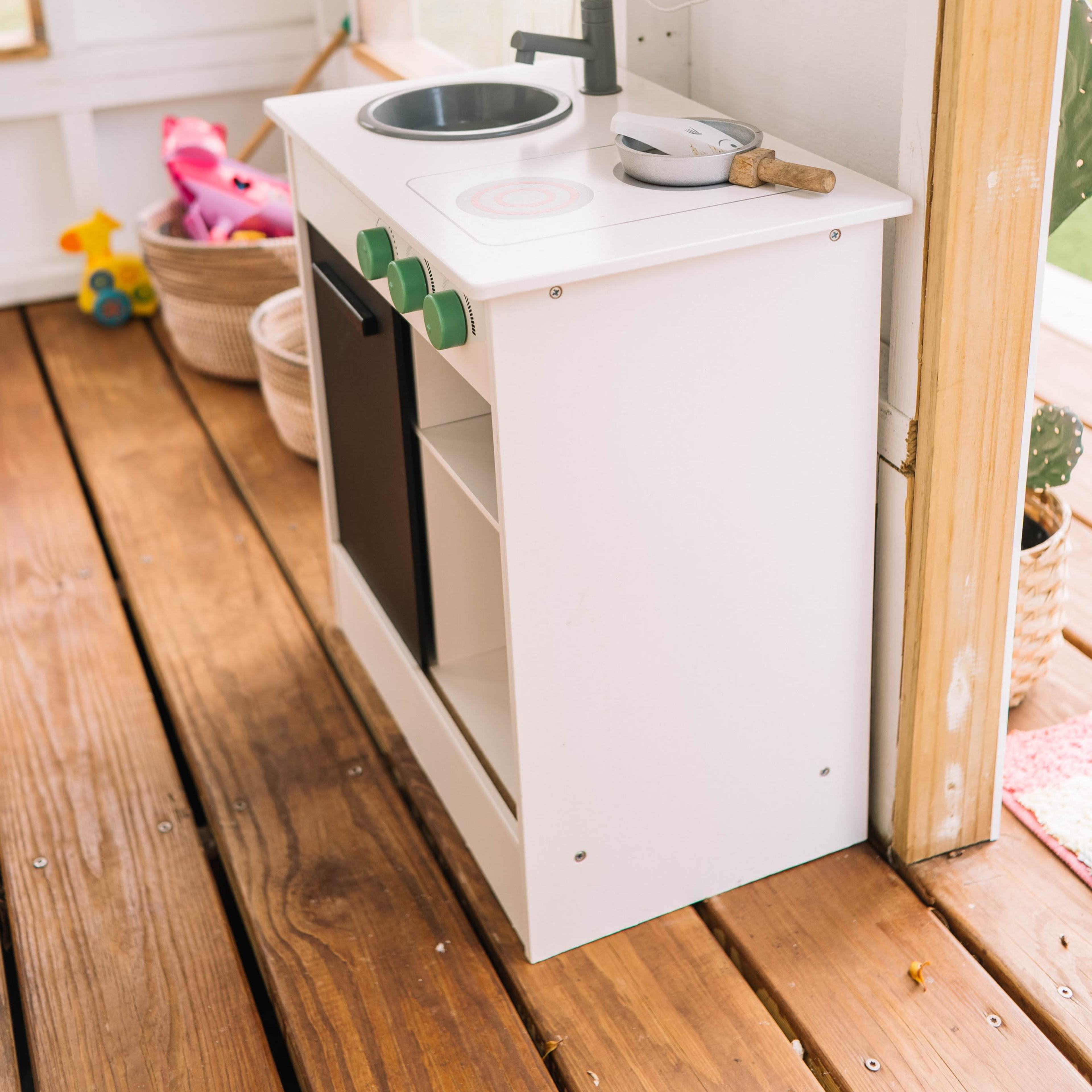 A small white play kitchen set with a sink, cooktop, and storage shelf, positioned on wooden flooring beside a window.