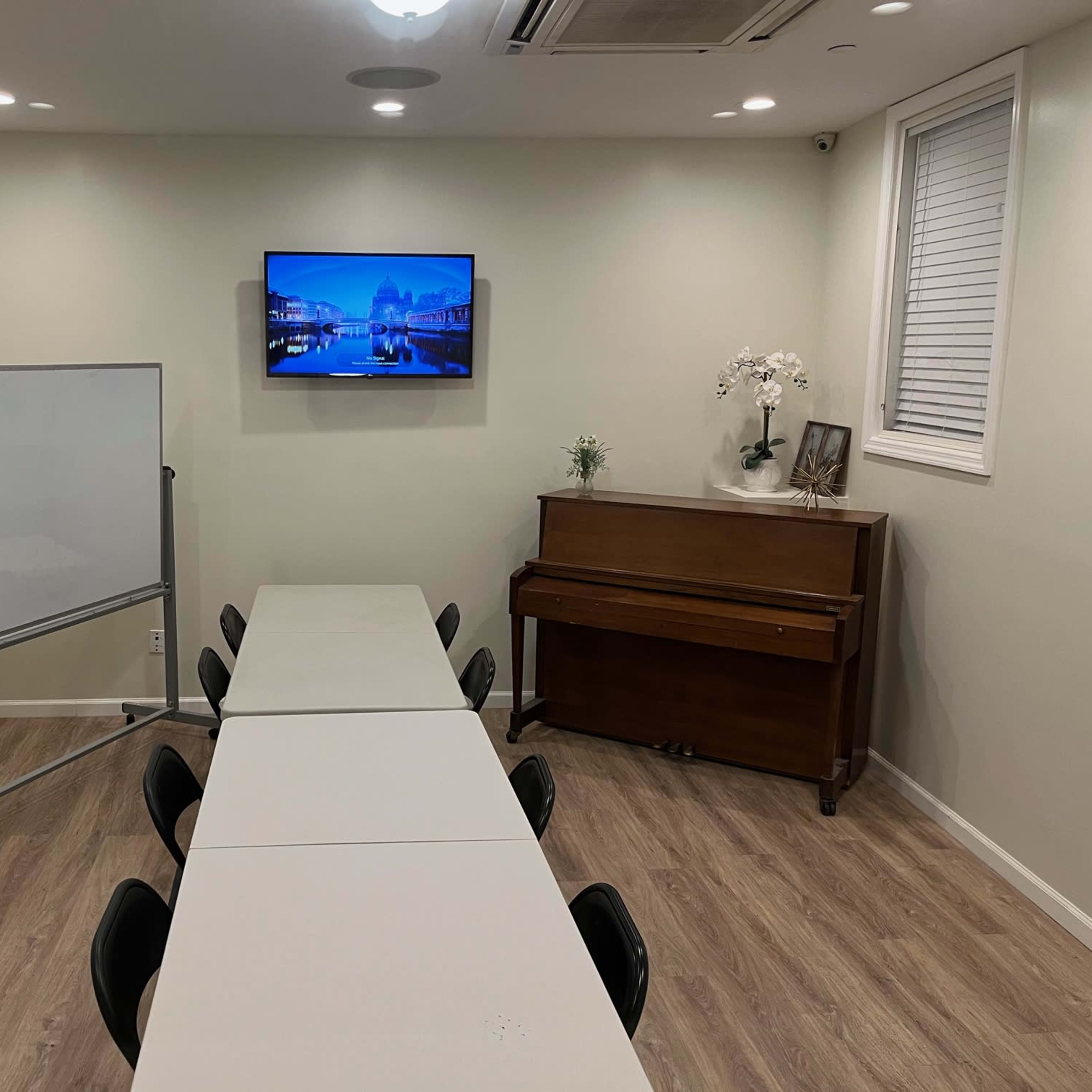 A small meeting room features a rectangular table with black chairs, a whiteboard on one side, and a piano opposite a wall-mounted TV.