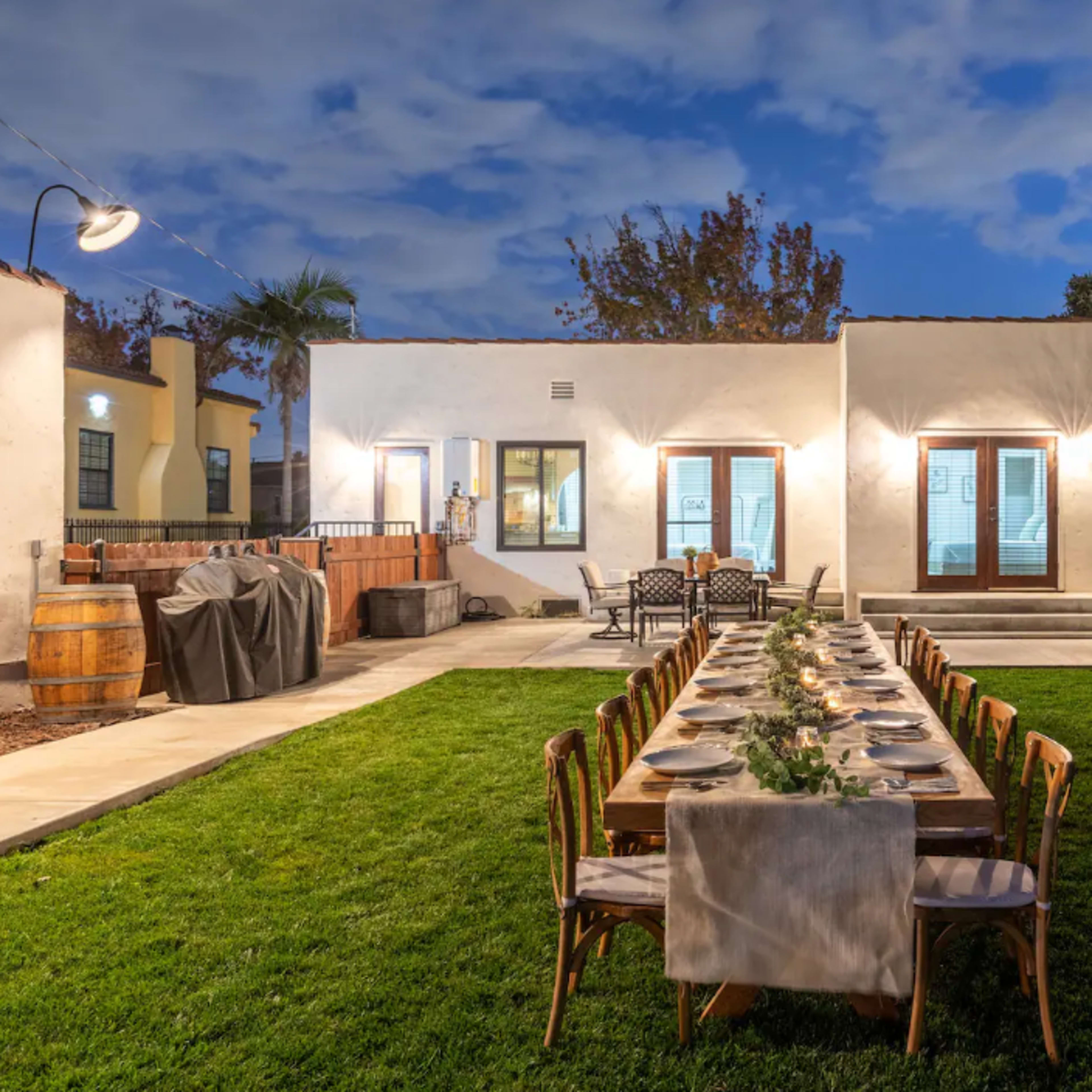 A long dining table is set up in a grassy yard behind a modern house, with chairs arranged around it and an orange tree in the background.
