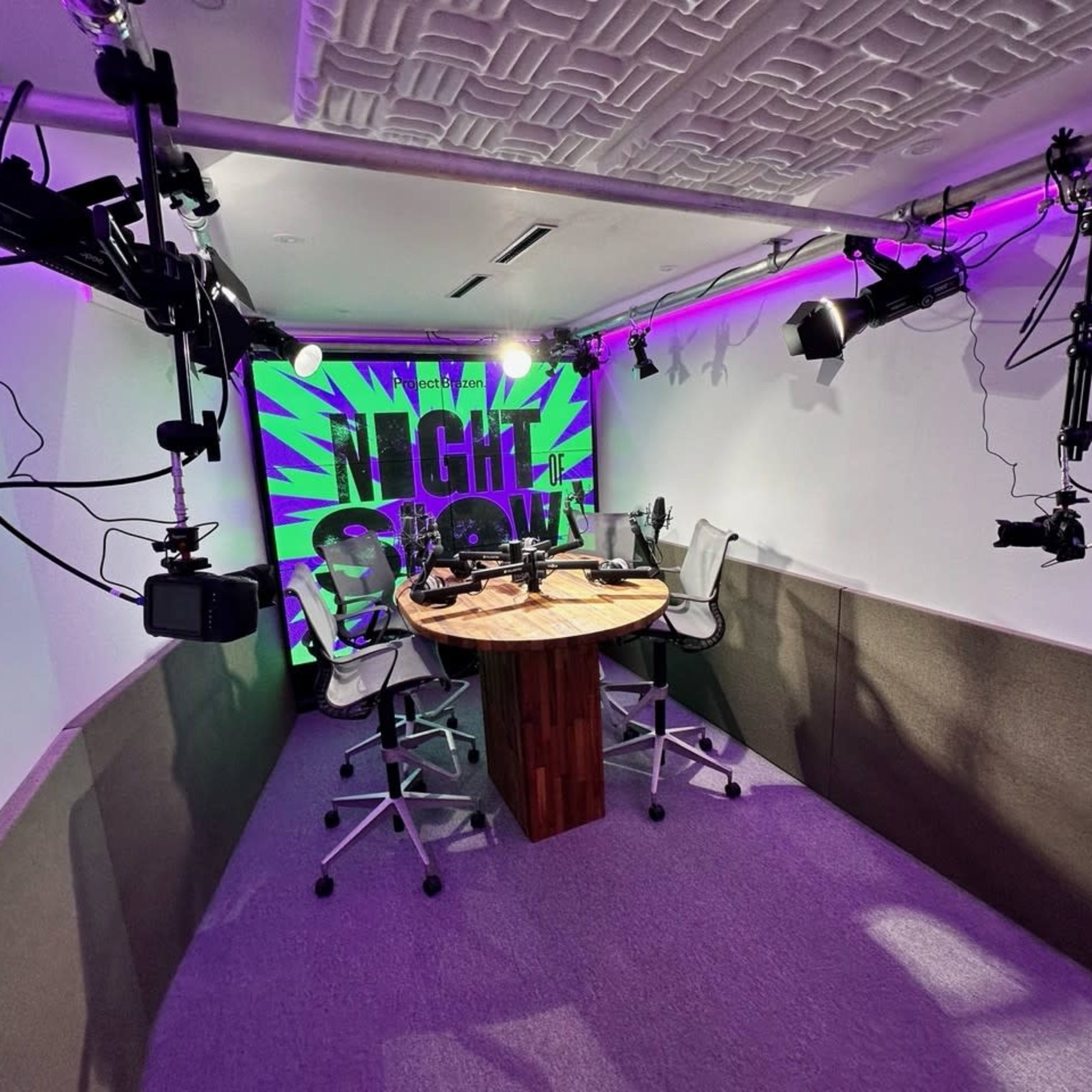 A podcast studio featuring a round table with chairs, surrounded by cameras and vibrant purple lighting on the walls.