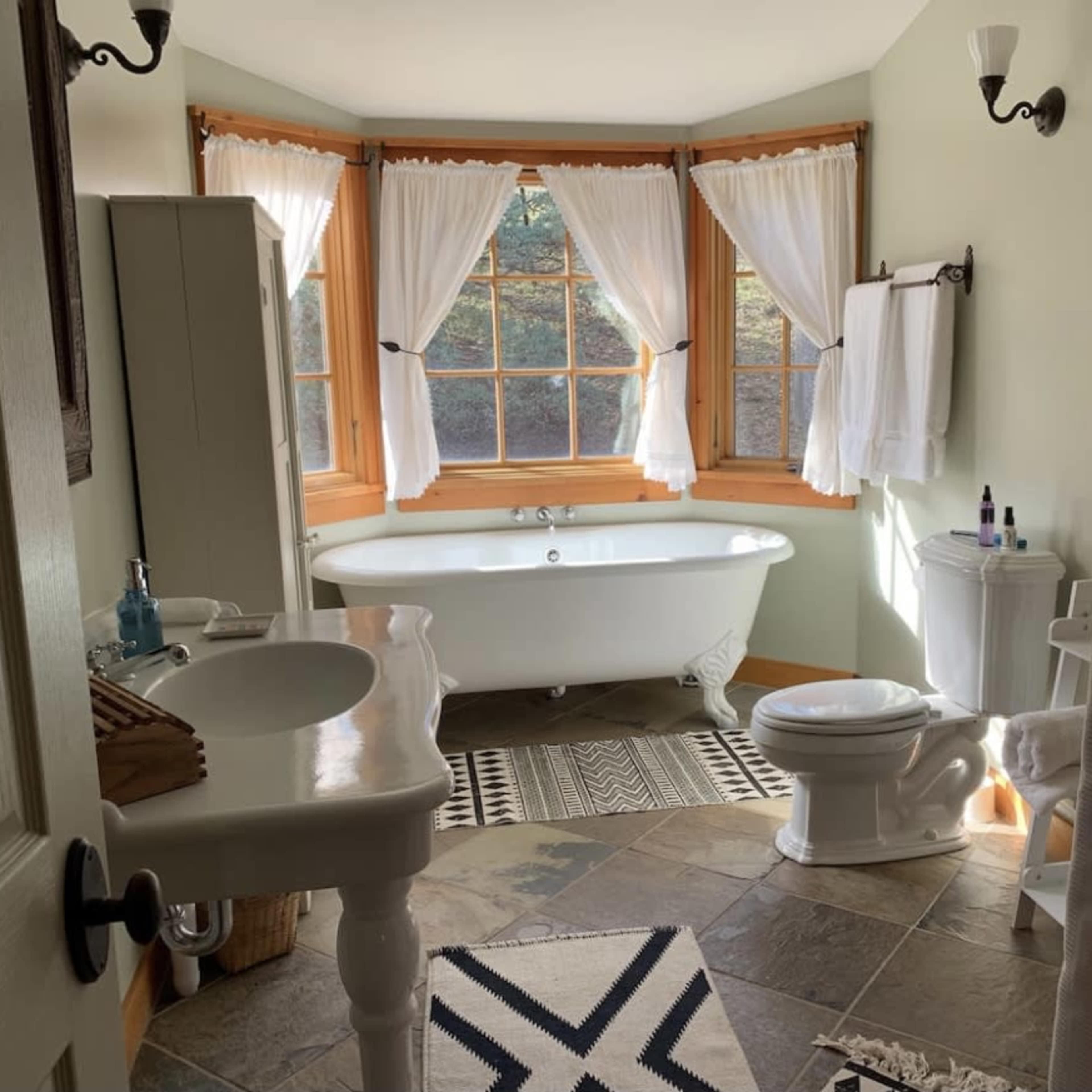 A bright bathroom features a freestanding tub positioned by large windows, a toilet, and a sink with a mirror, all surrounded by decorative tiles and a patterned rug.