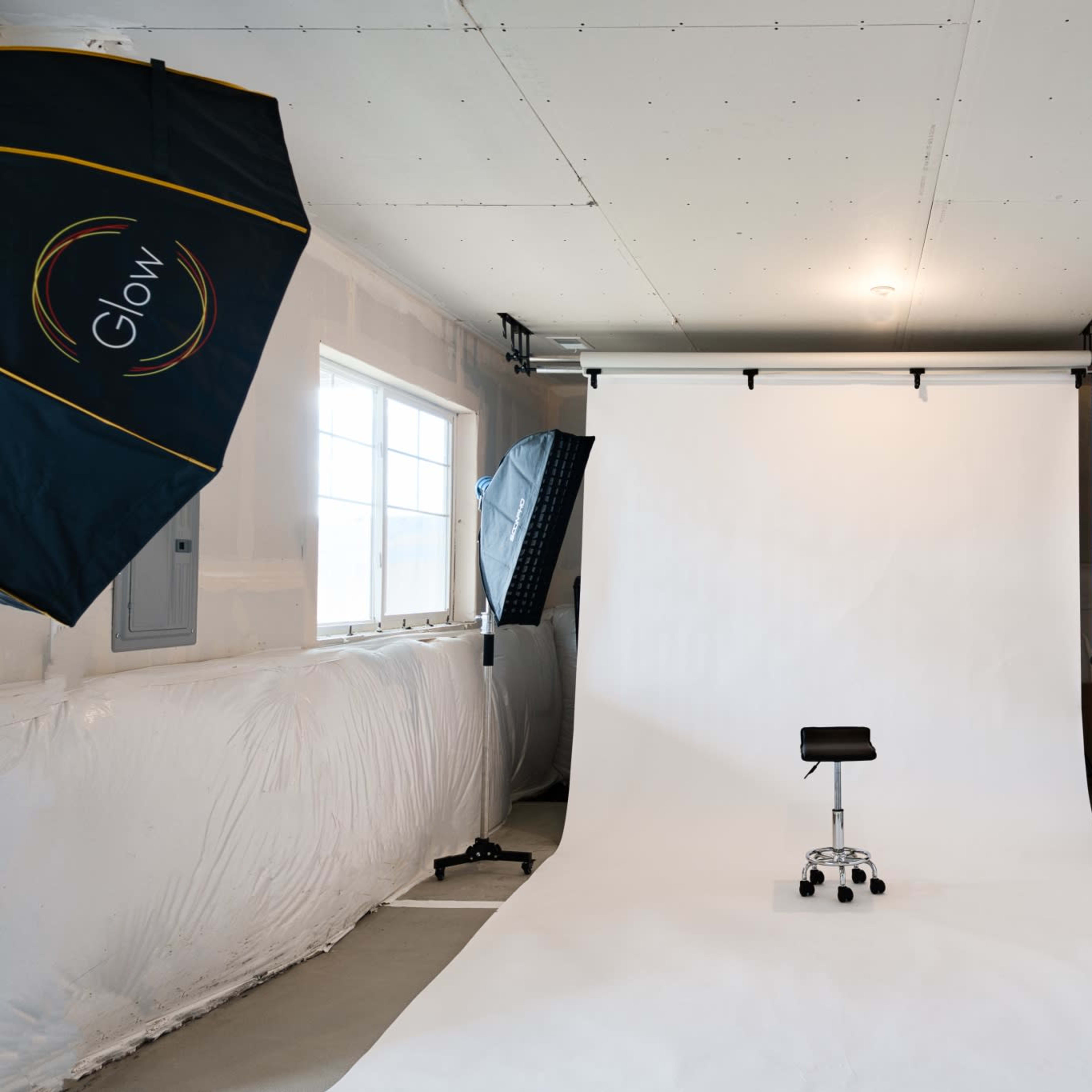 A photography studio is set up with a white backdrop, a stool in the center, and various lighting equipment positioned around the room.
