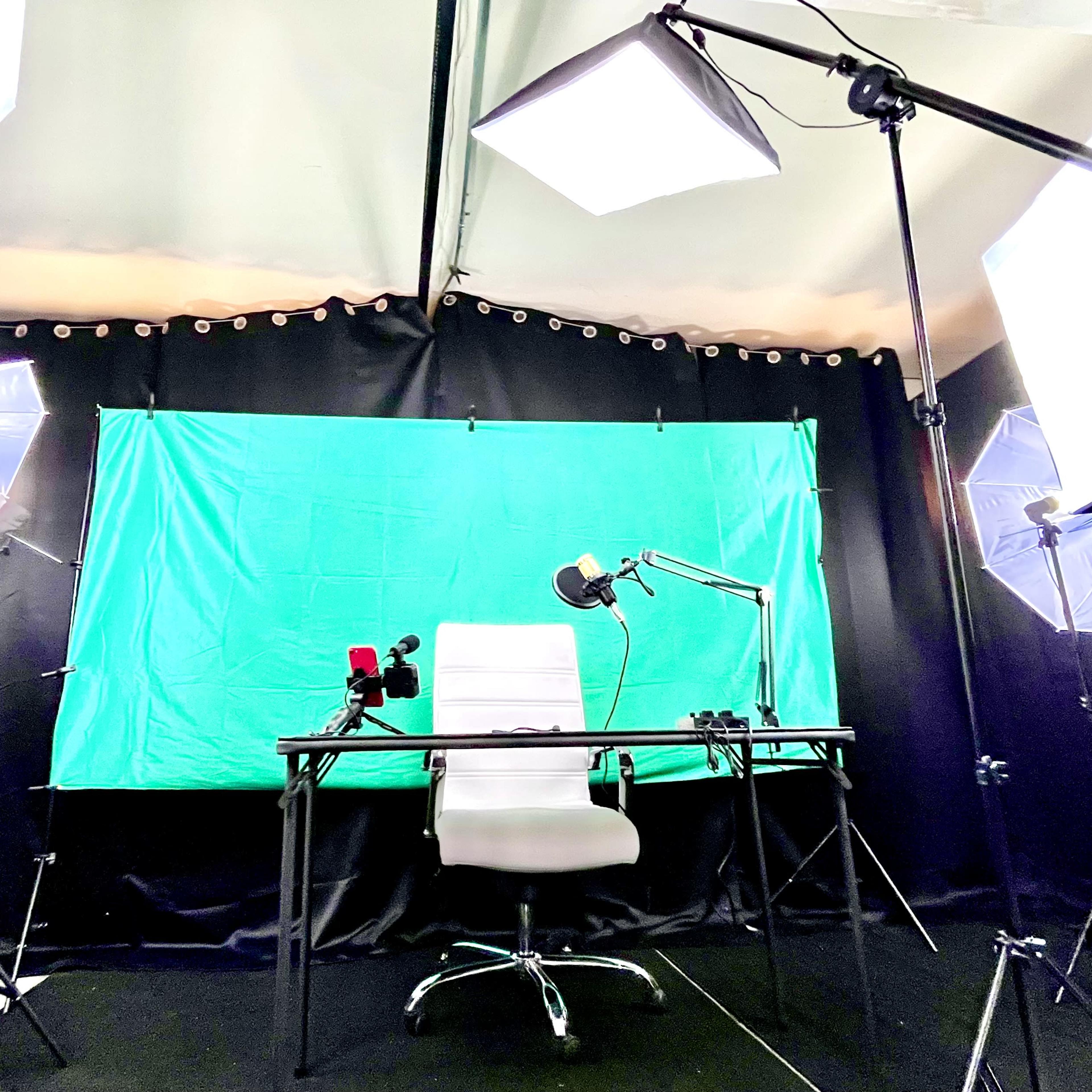 A recording studio setup with a green backdrop, a white chair, a desk, and several lighting devices positioned around the space.