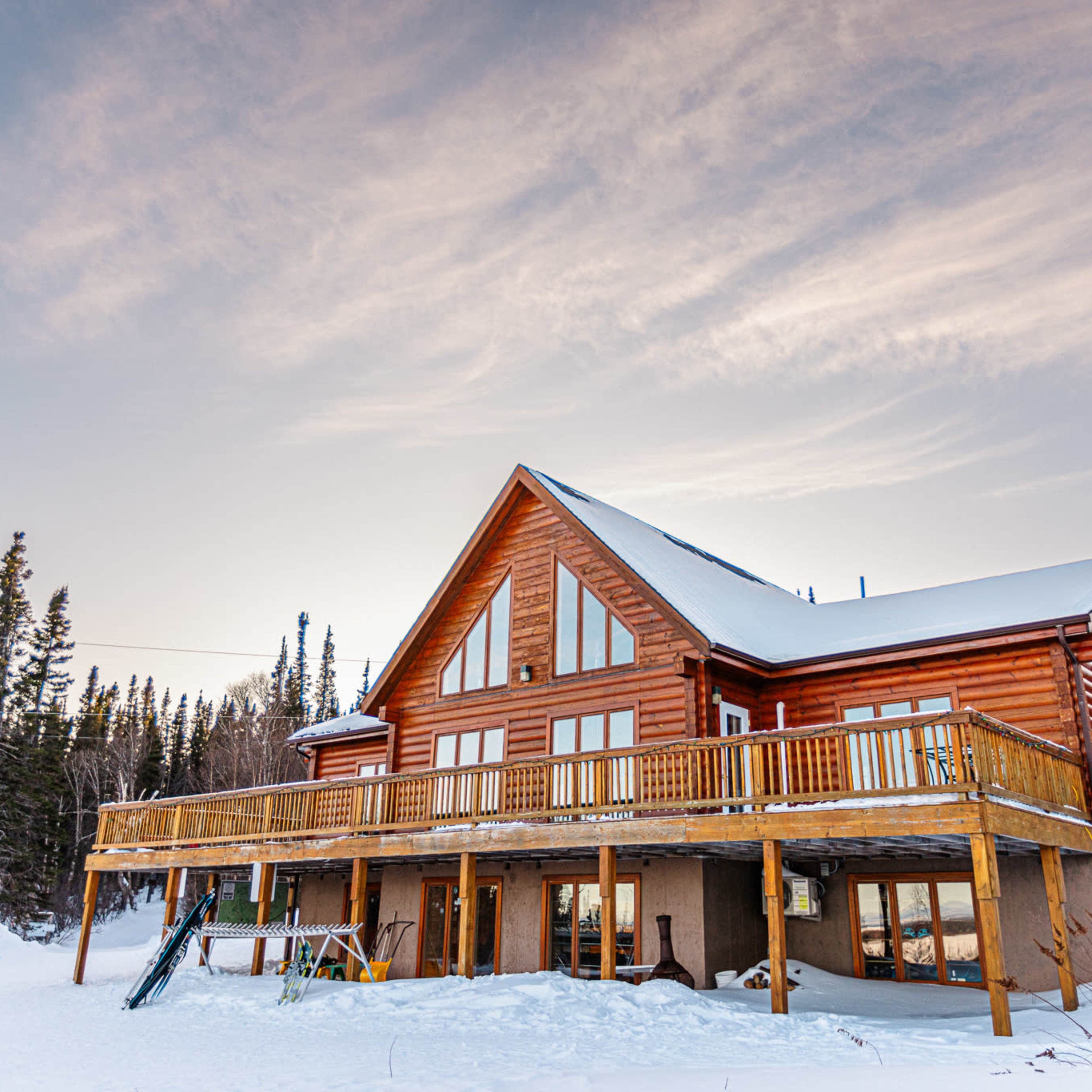 A large wooden cabin with a wraparound deck and expansive windows is situated in a snowy landscape surrounded by trees.