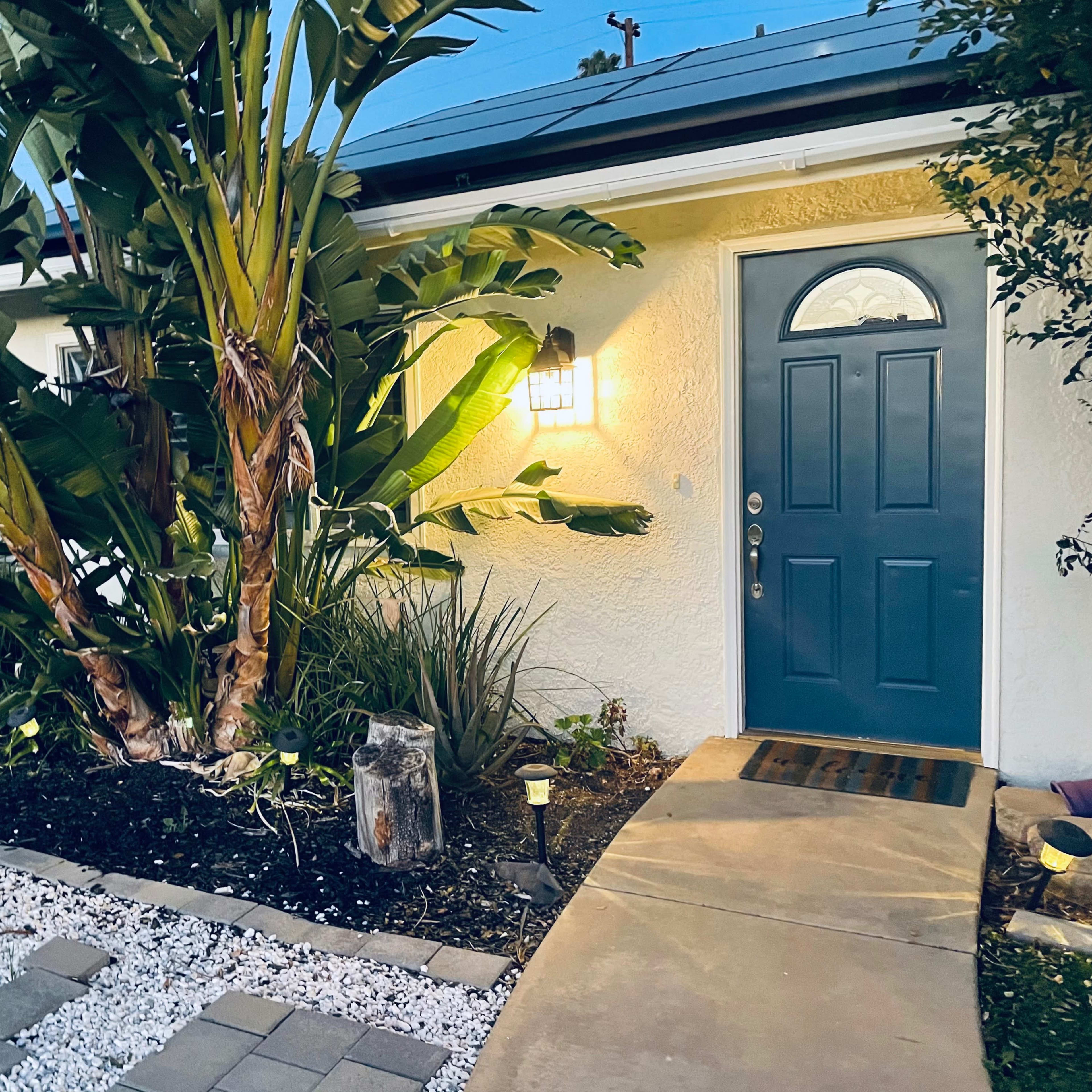 A brightly lit front door is flanked by tall tropical plants and a pathway made of concrete slabs and white gravel.