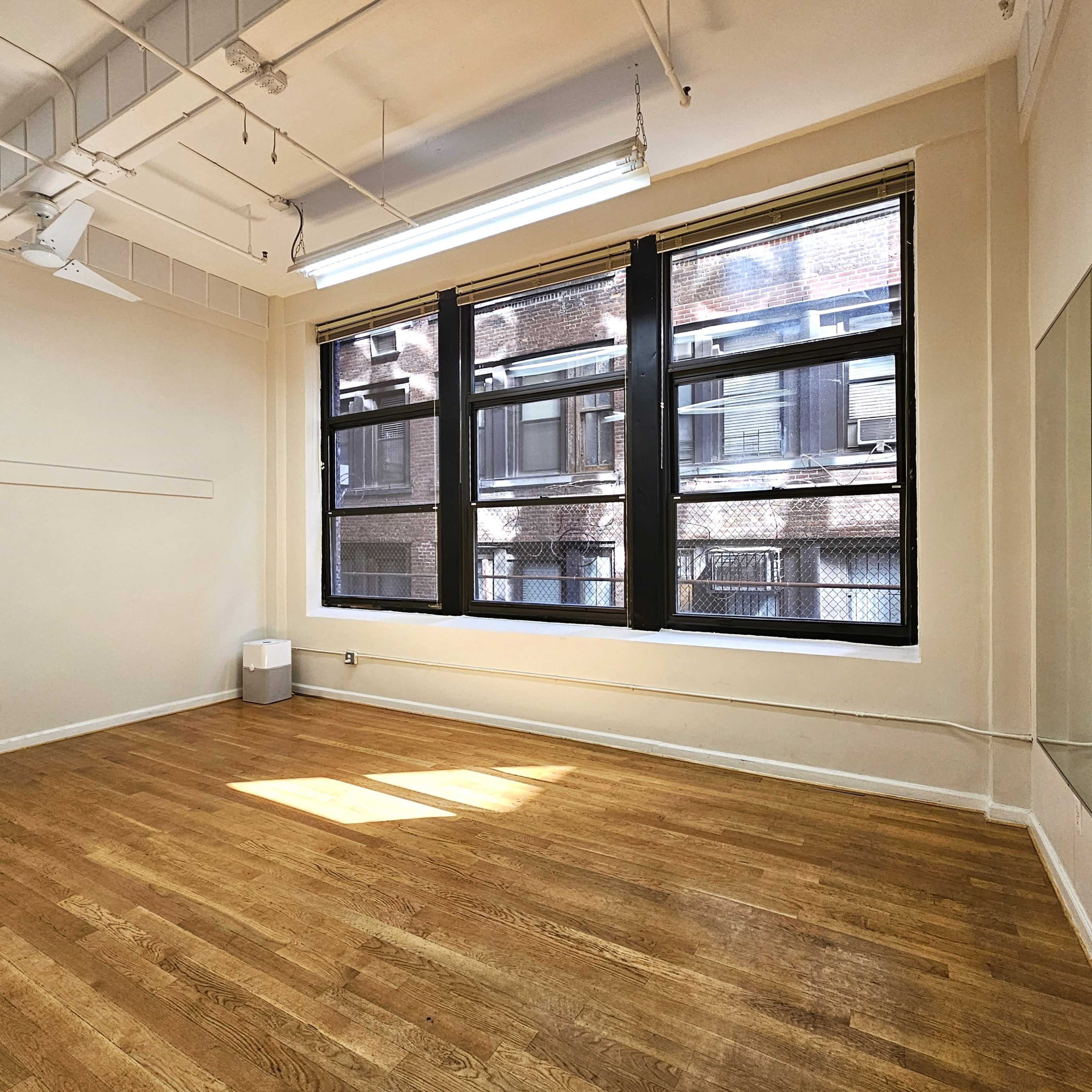 A spacious room with wooden floors, large windows, and a mirror on one wall.