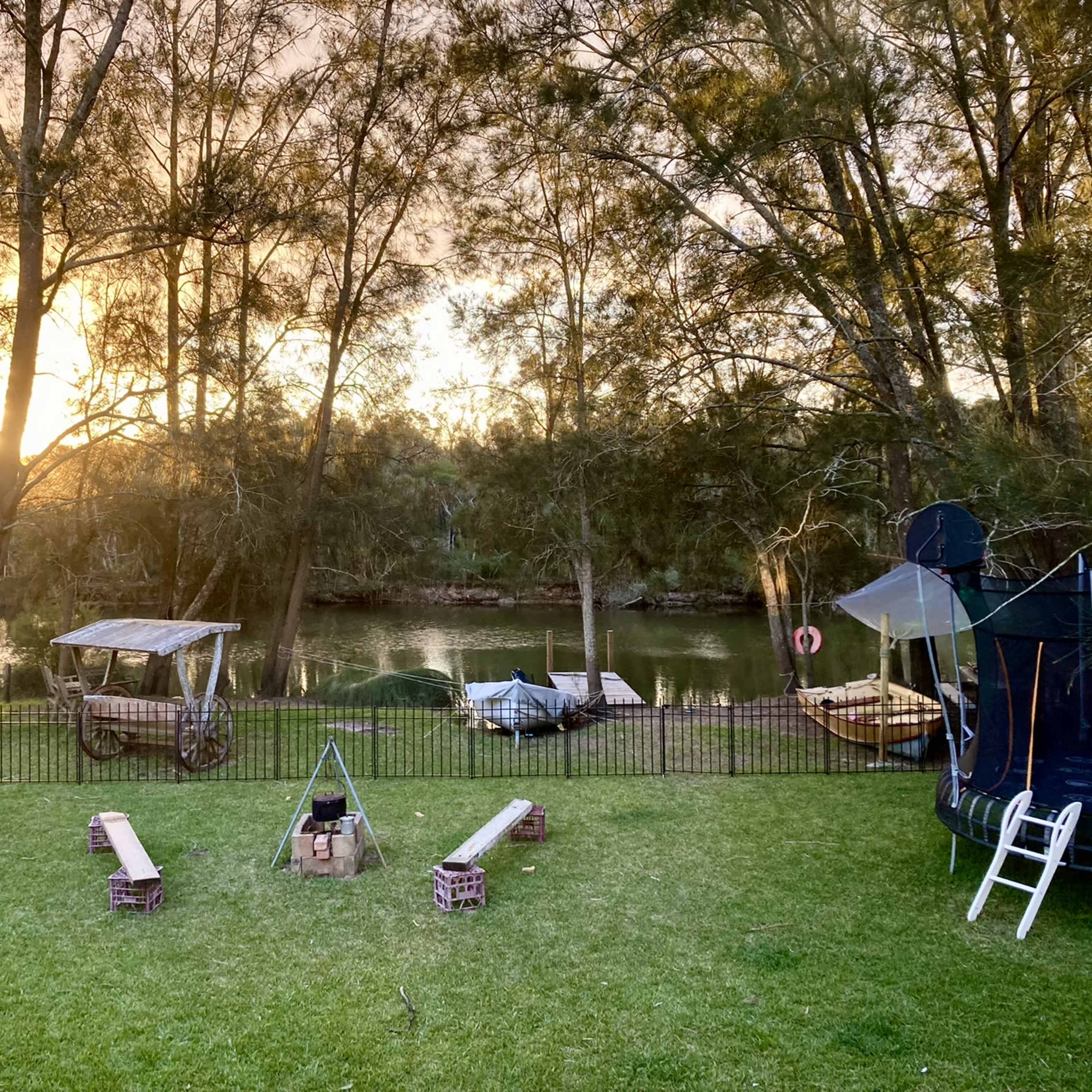 The scene features a grassy area by a river with a trampoline, wooden benches arranged around a fire pit, and several boats moored nearby under tall trees.