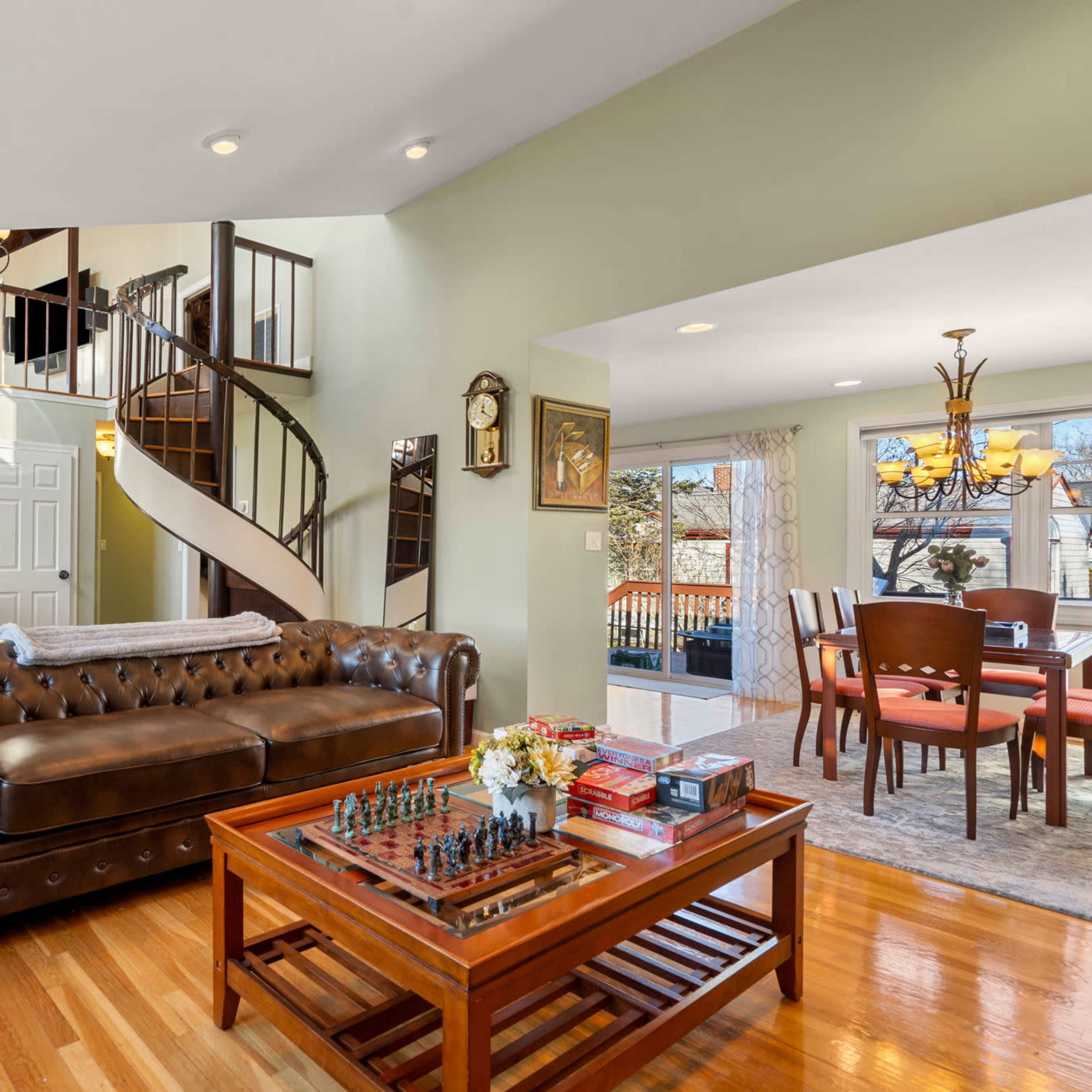 A living room with a leather chesterfield sofa, a wooden coffee table with a chess set on it, and a spiral staircase leading to the upper floor.