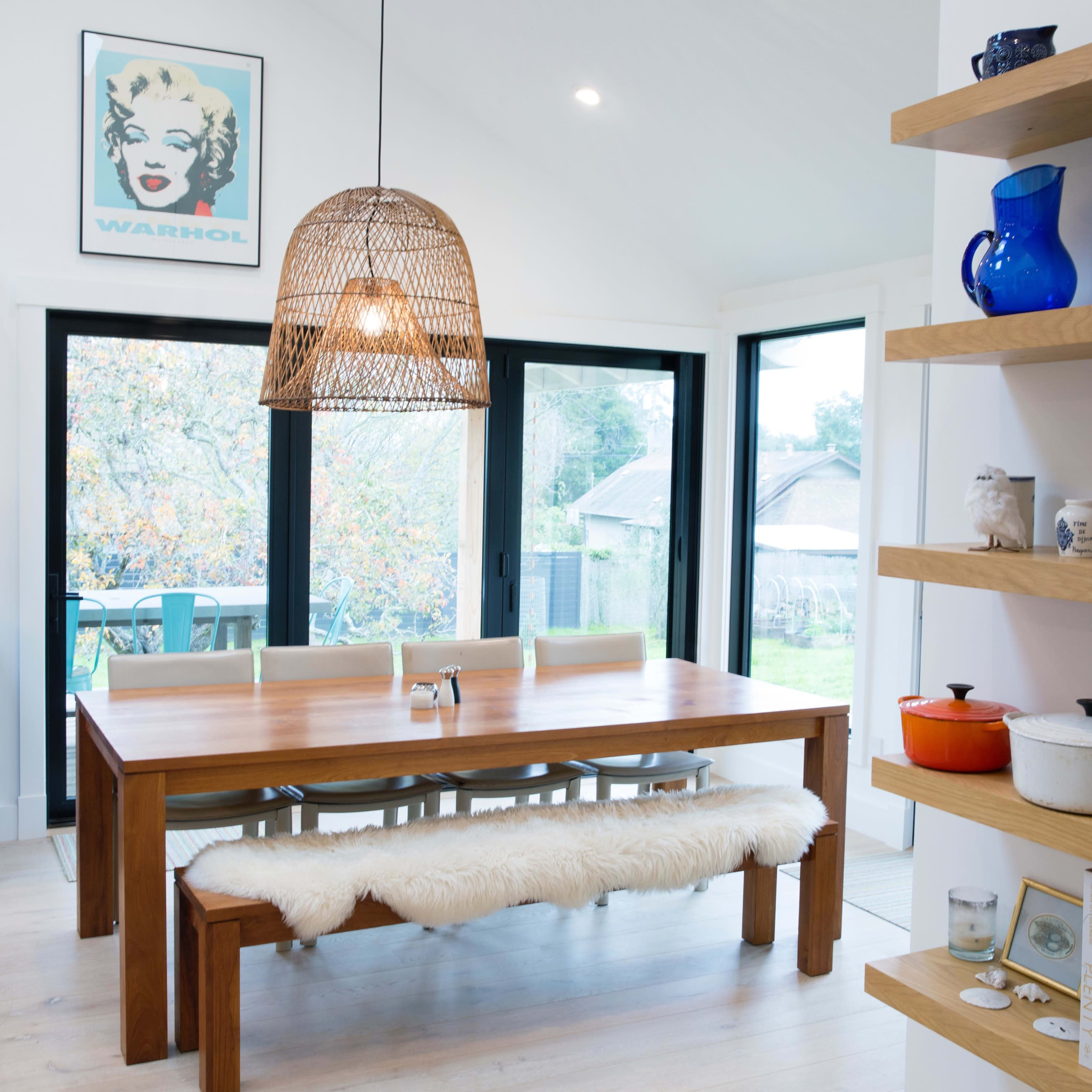 A modern dining area featuring a wooden table with a bench and chairs, pendant lighting, and shelves displaying various dishes and decorative items.