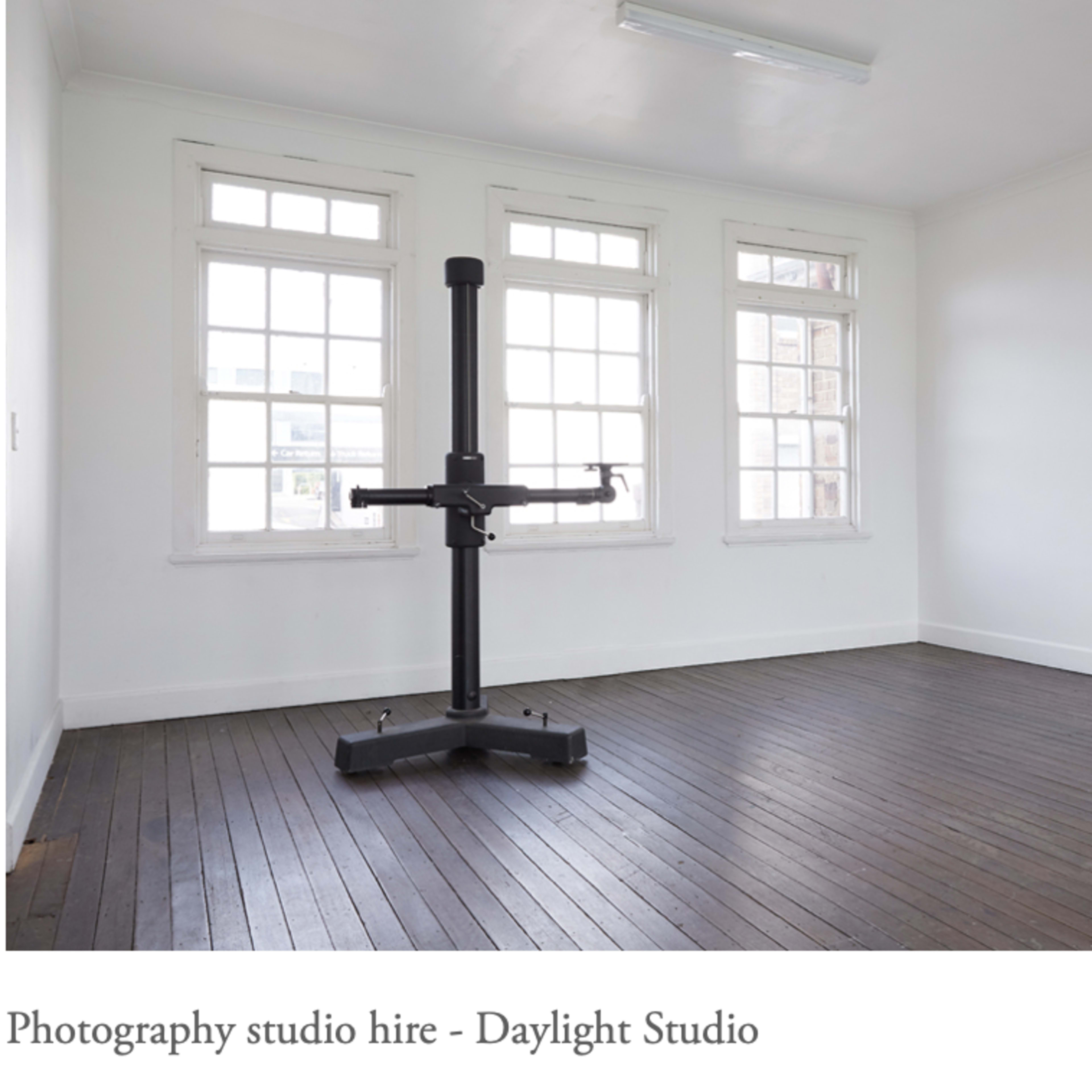 A minimally furnished photography studio with large windows and a central mounting stand on a wooden floor.