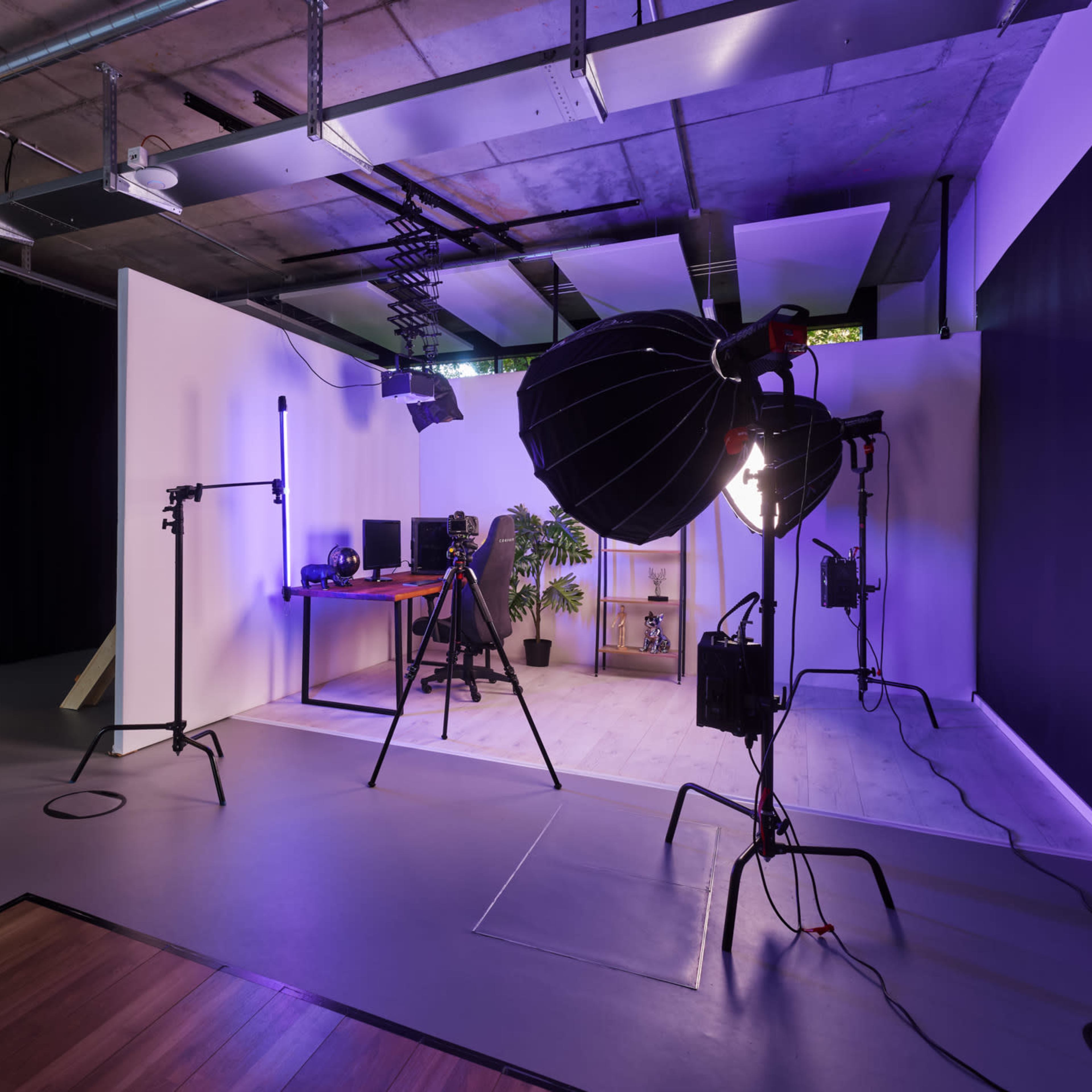 A modern photography studio featuring various lighting equipment, a work desk with computers, and a large potted plant.