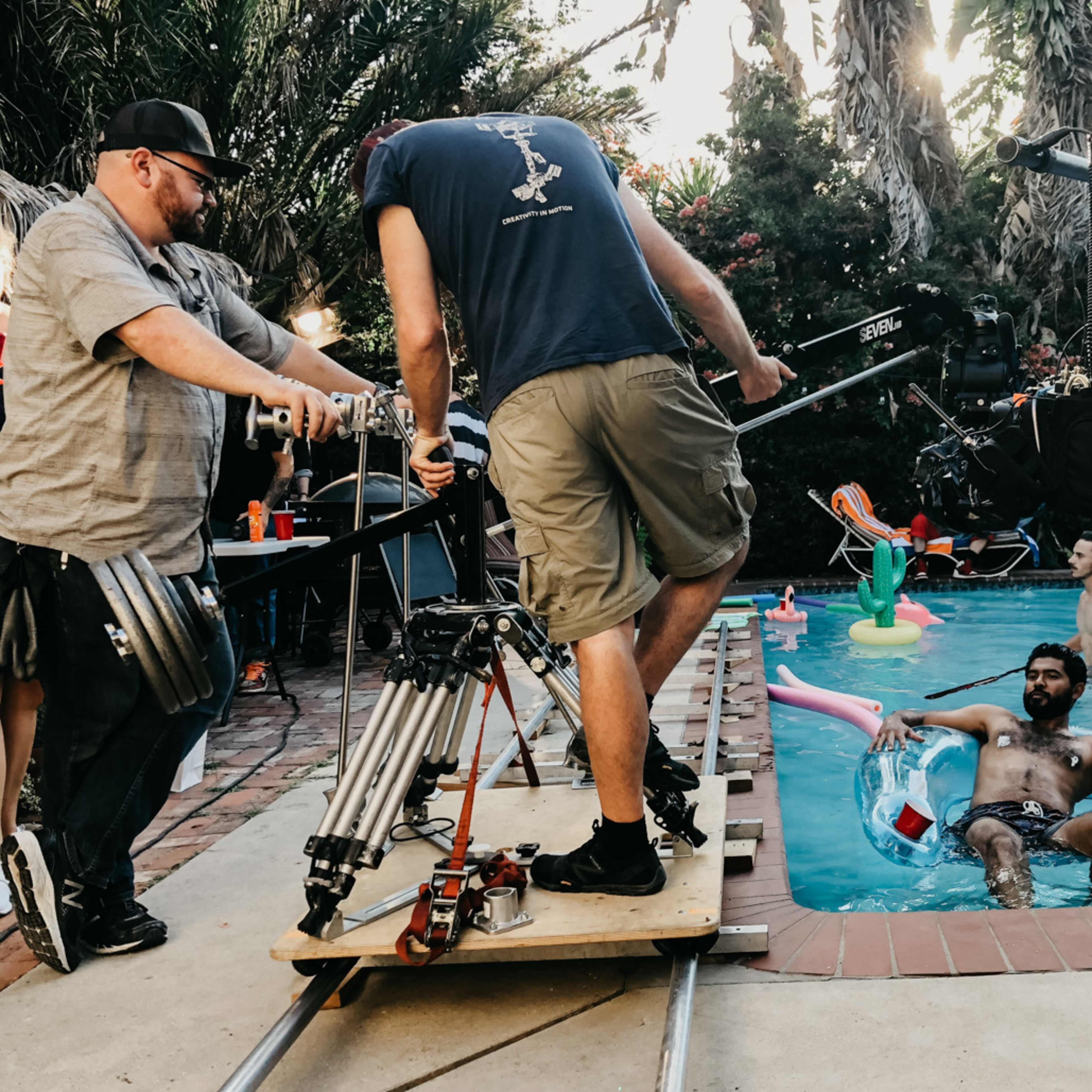A film crew is set up by a swimming pool, with one person sitting on an inflatable float while another operates a camera on a dolly.
